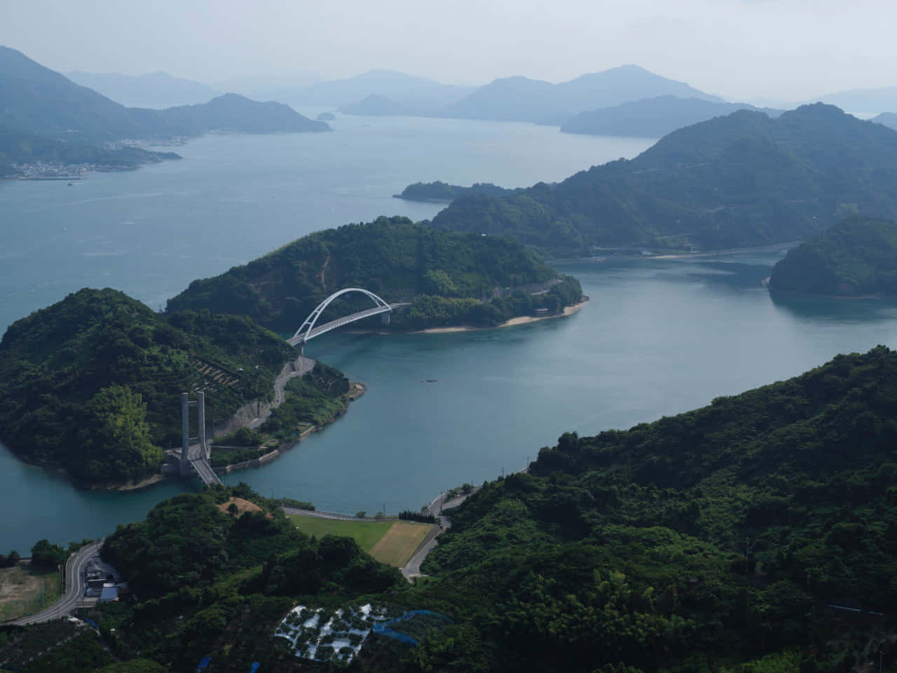  壁紙 — 廣闊的鳥瞰圖，一座現代拱橋橫跨寧靜的藍色海灣，周圍是鬱鬱蔥蔥的綠色山脈。