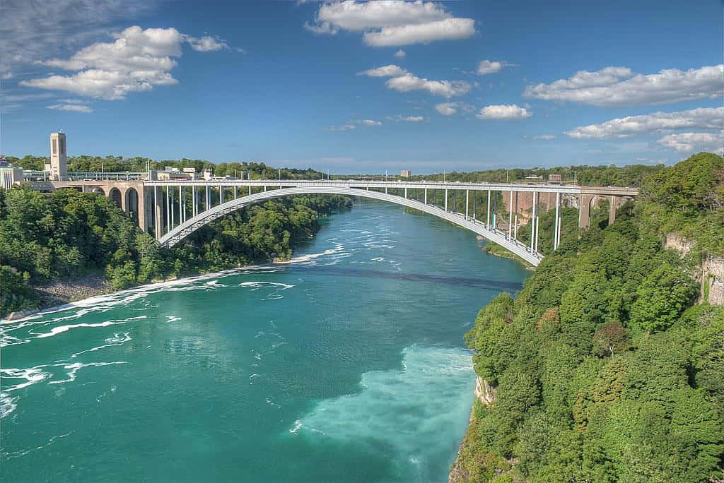 Fall Bridge 桌布 壁紙 — 尼亞加拉大橋橫跨碧綠河流的廣闊航拍視圖，兩岸綠樹成蔭，天空湛藍，白雲點綴。