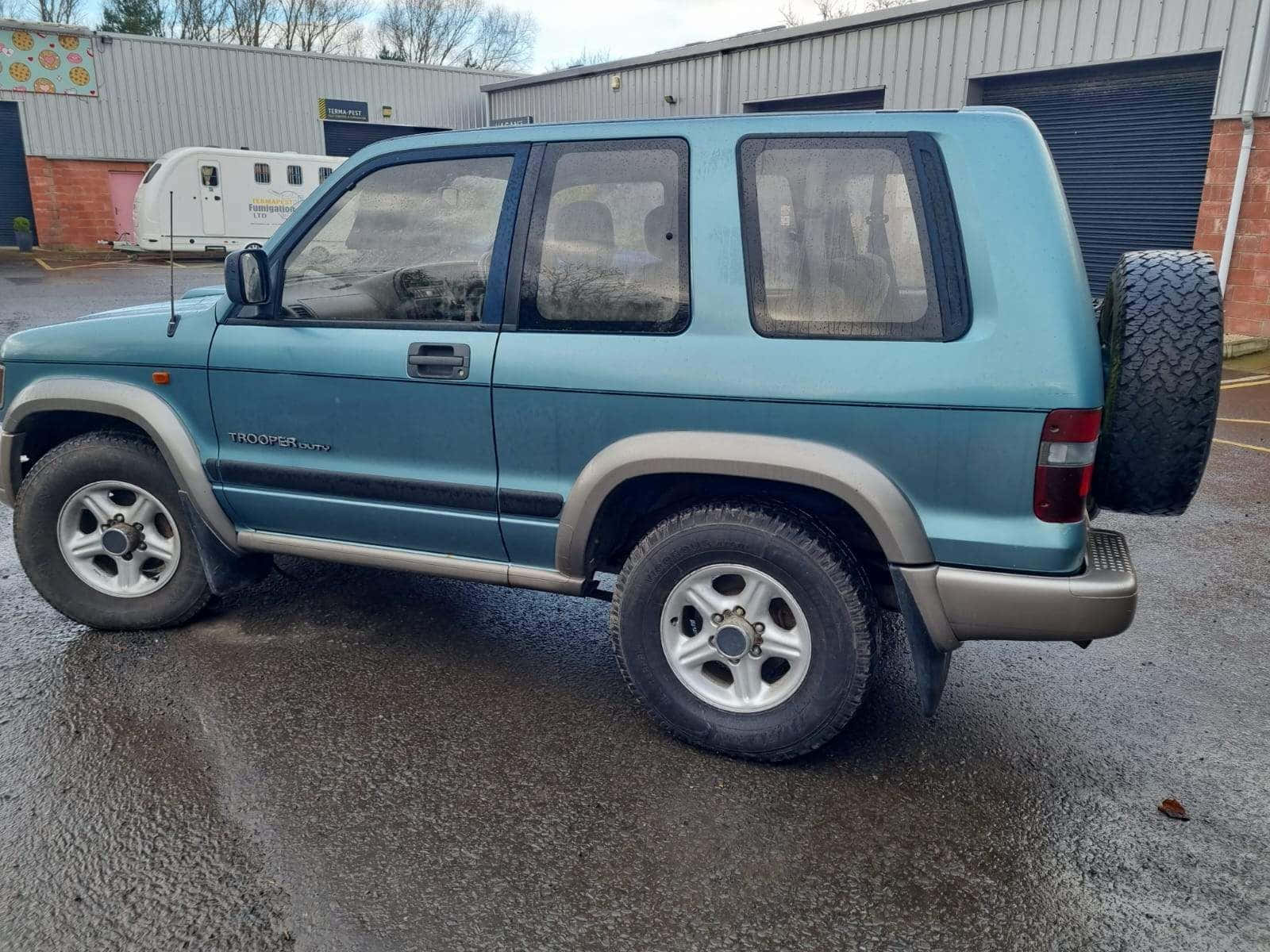  壁紙 — 一輛青綠色鈴木Jimny Trooper SUV停放在工業建築物前的濕路面上，天空多雲。