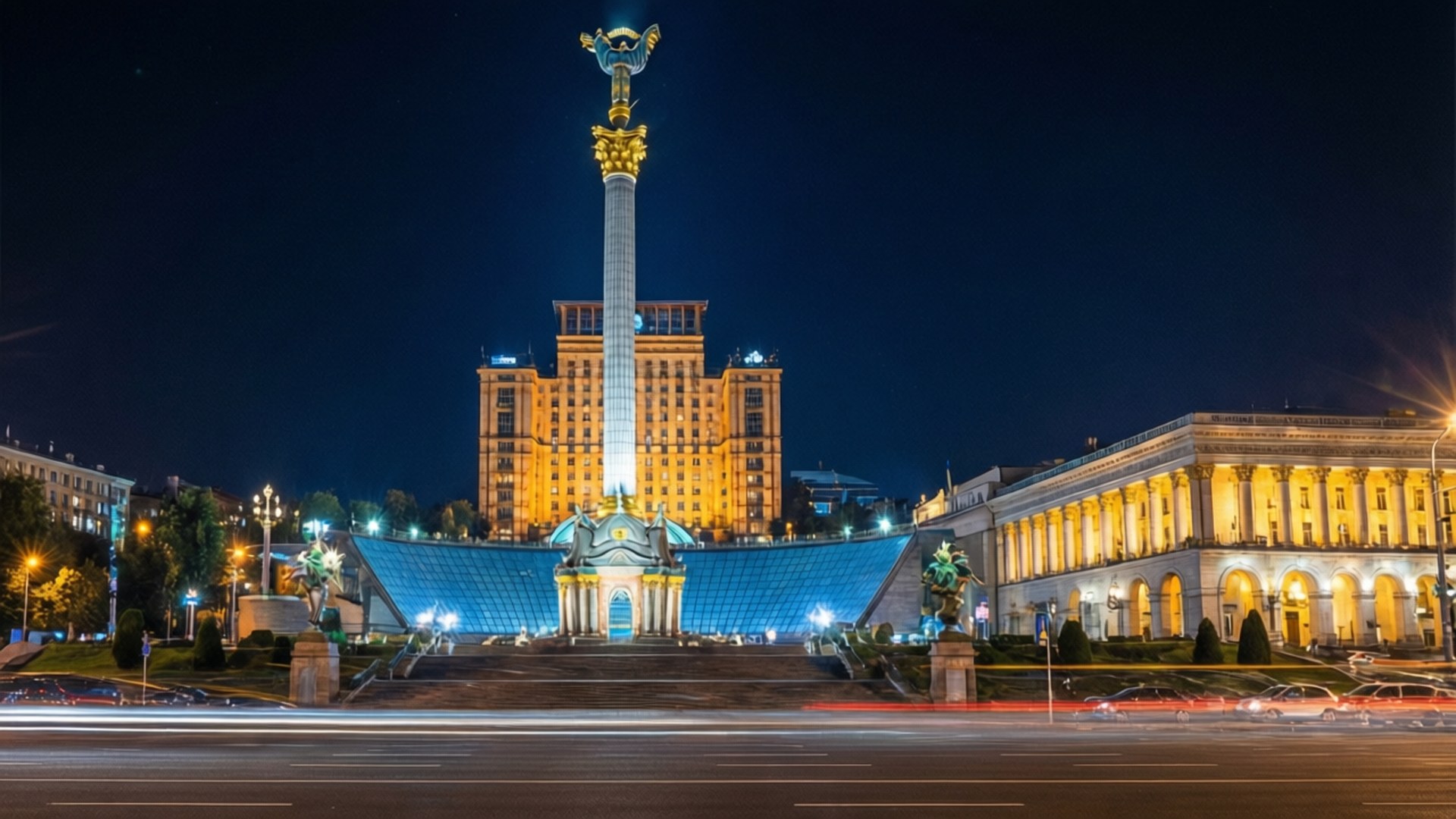 Kyiv Night Lights 壁紙 — 一座高聳的紀念碑頂端有金色雕像，在夜晚照亮了廣場，兩旁是宏偉的建築，車燈劃過形成光軌。