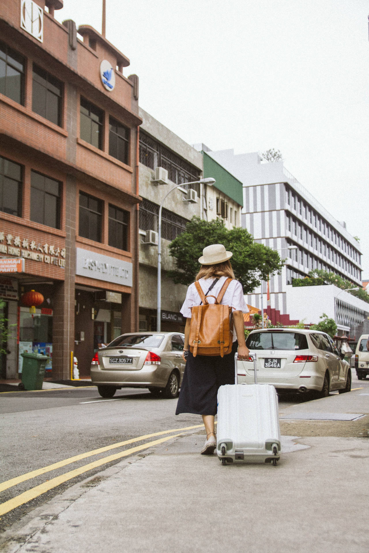 一個戴著帽子和背包的人，拖著行李箱穿過城市街道。 — 4K 壁紙