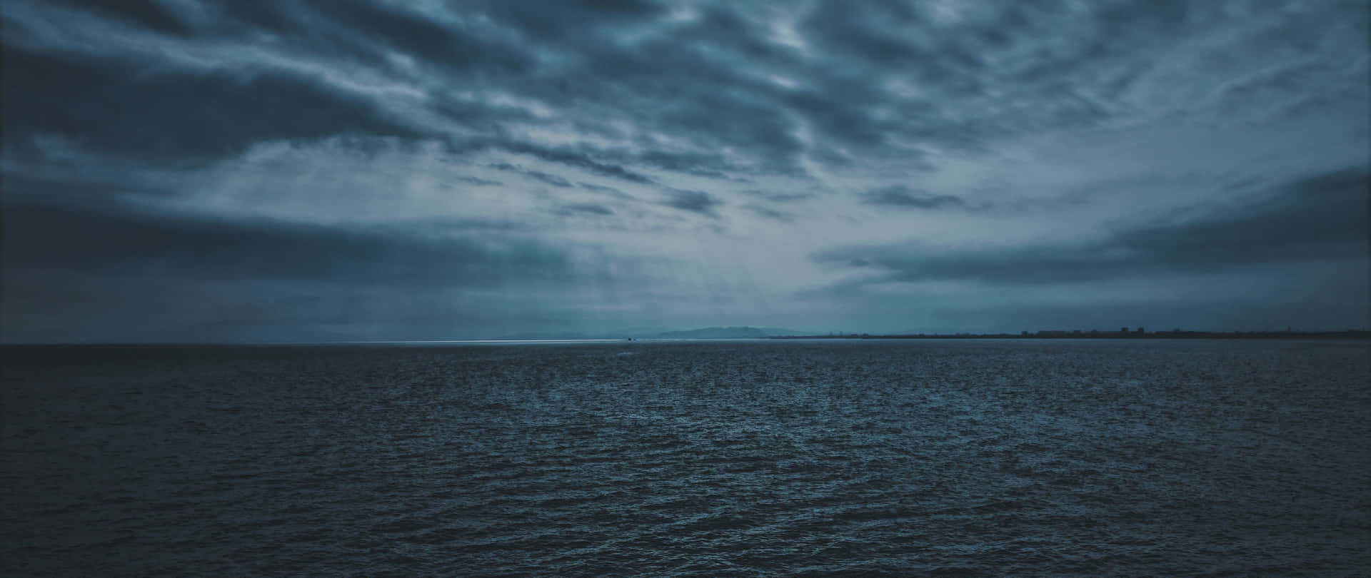  壁紙 — 廣闊的黑暗海洋，天空陰沉，雲層戲劇性，地平線上有遙遠的陸地。