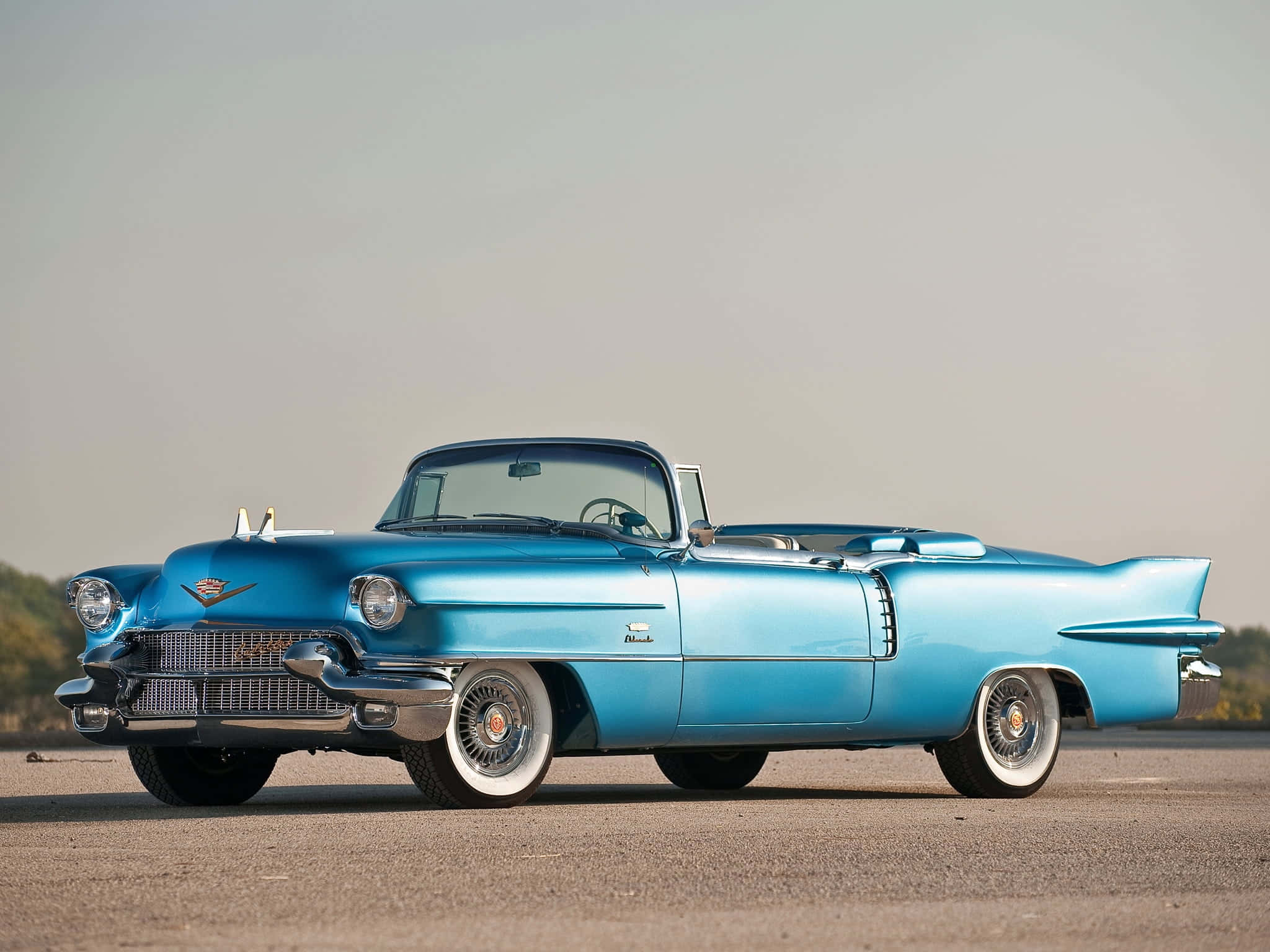 Cadillac Eldorado 桌布 壁紙 — 一輛鮮藍色的1959年Cadillac敞篷車在柔和的日光下，在一個有遠處樹木的開放鋪砌場地上閃閃發光。
