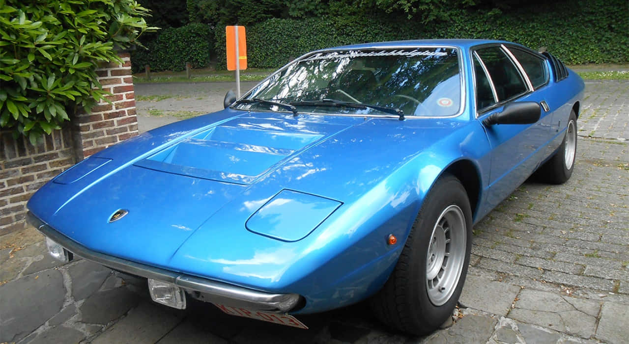 Lamborghini Urraco 桌布 壁紙 — 一輛充滿活力的藍色經典藍寶堅尼Countach停放在石頭車道上，旁邊有綠植和磚牆。