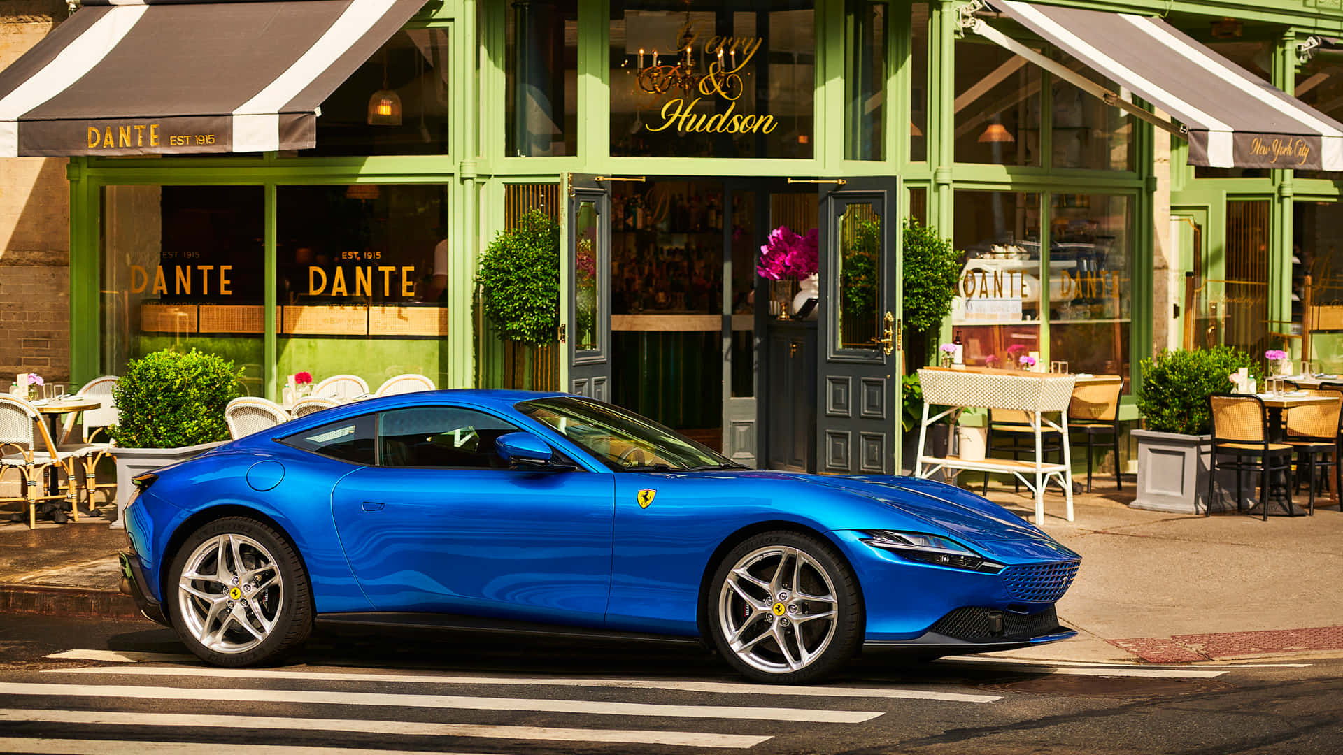 Ferrari Roma 桌布 壁紙 — 一輛鮮藍色的法拉利 Roma 停放在陽光明媚的街道上，前面是綠色店面，設有戶外咖啡座。