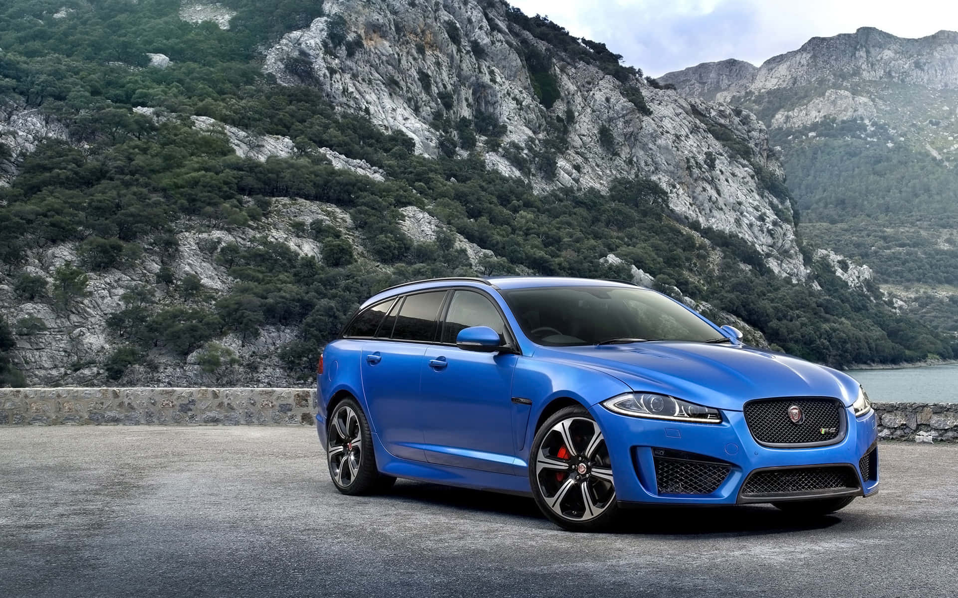 Jaguar Xfr 桌布 壁紙 — 一輛充滿活力的藍色積架旅行車停泊在沿海公路上，背景是崎嶇的山脈和綠意。