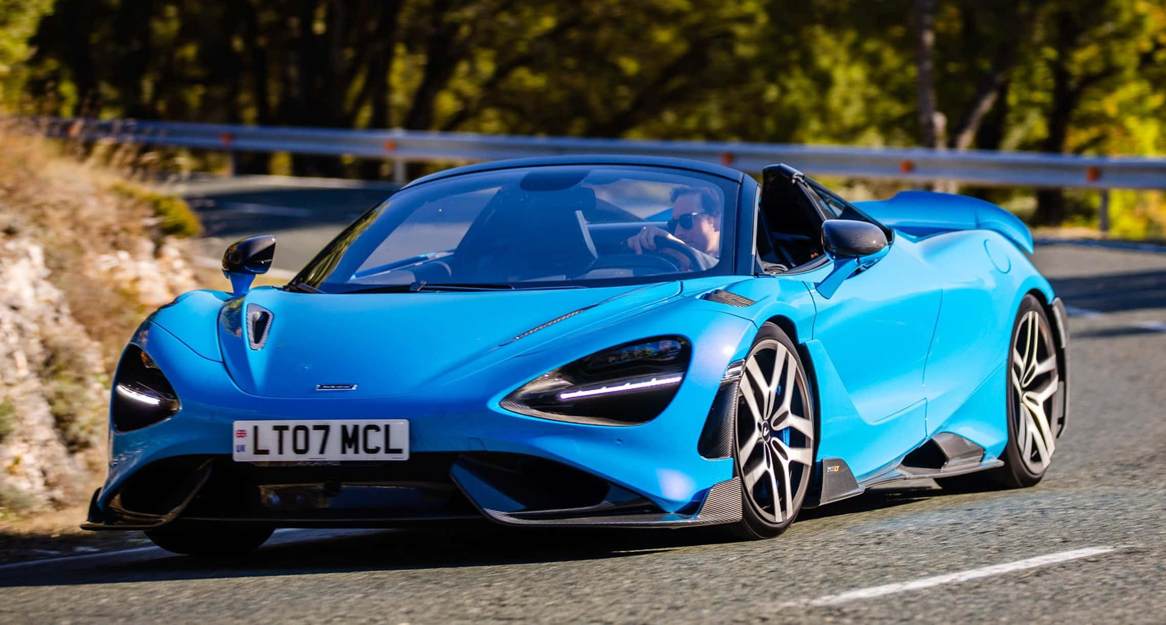 Mclaren 765lt 桌布 壁紙 — 一輛充滿活力的藍色McLaren 720S Spider在陽光明媚的山路上疾馳，背景是茂密的綠色樹木。