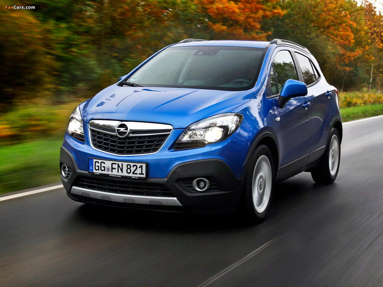 Opel Mokka 桌布 壁紙 — 一輛充滿活力的藍色 Opel Mokka SUV 在道路上疾馳，背景中秋天的樹木模糊不清。
