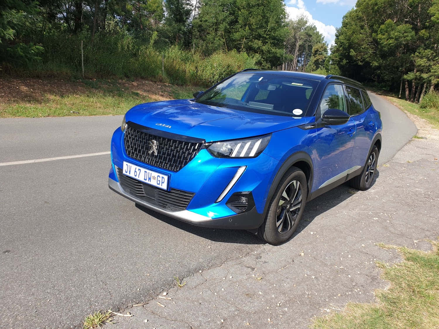 Peugeot 2008 桌布 壁紙 — 一輛充滿活力的藍色標緻SUV停泊在蜿蜒的瀝青路上，周圍是茂密的綠樹，天空明亮晴朗。