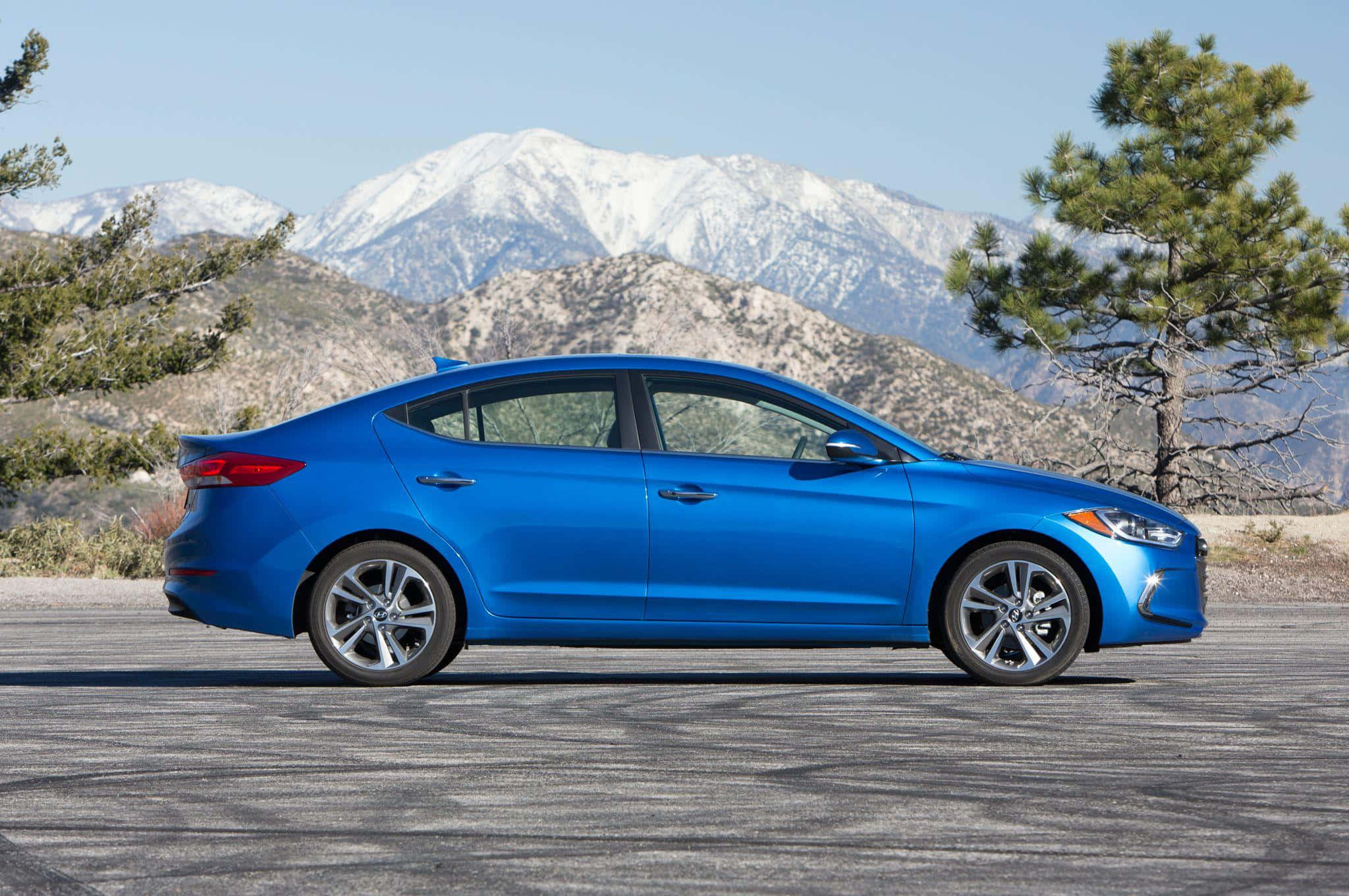 Hyundai Elantra 桌布 壁紙 — 一輛充滿活力的藍色轎車停在鋪砌的道路上，背景是白雪覆蓋的山脈和松樹，天空清澈湛藍。