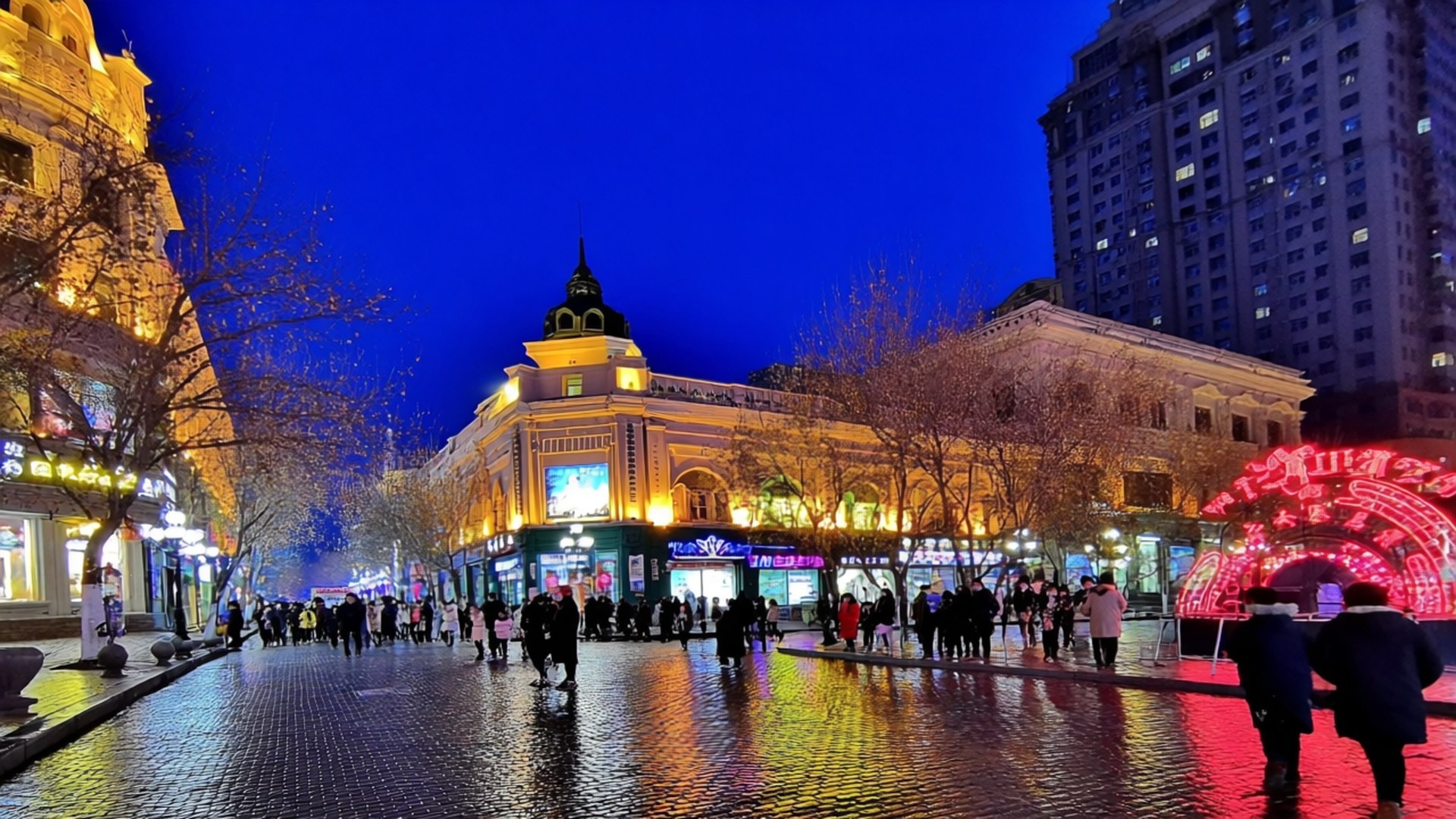 Central Street Neon Nights 壁紙 — 黃昏時分充滿活力的城市街道，金色燈光照亮的歷史建築倒映在濕漉漉的鵝卵石上，人們在發光的紅色拱門附近行走。