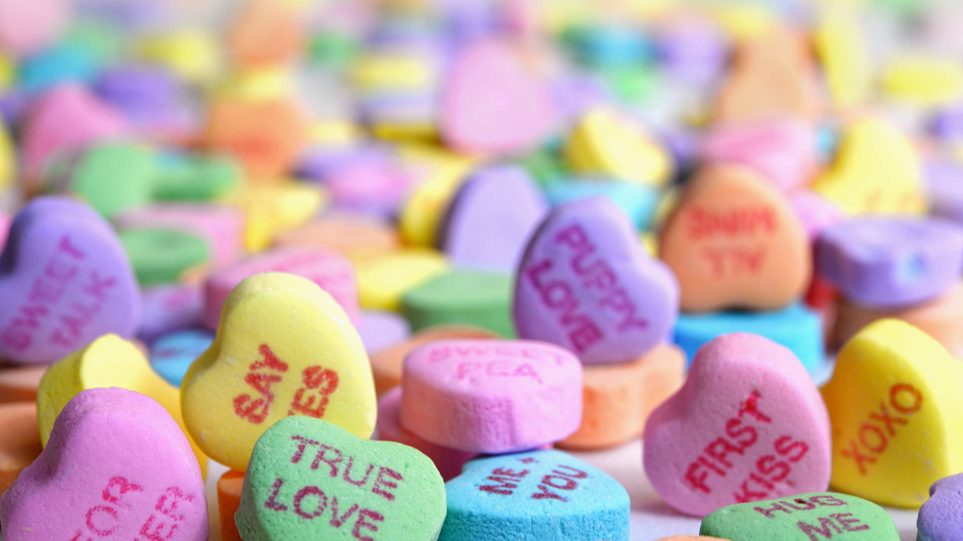 一堆色彩繽紛的心形糖果，上面有甜蜜訊息如 true love 和 puppy love。 — Pastel 壁紙
