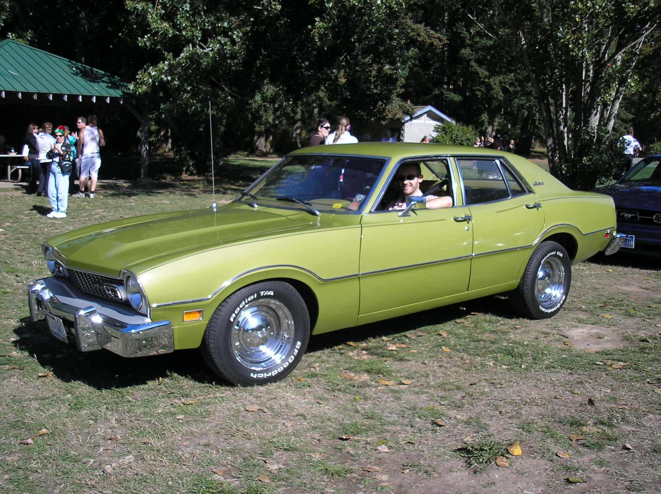 Ford Maverick 桌布 壁紙 — 一輛充滿活力的萊姆綠色經典汽車停在草地上，人們聚集在附近的樹下。