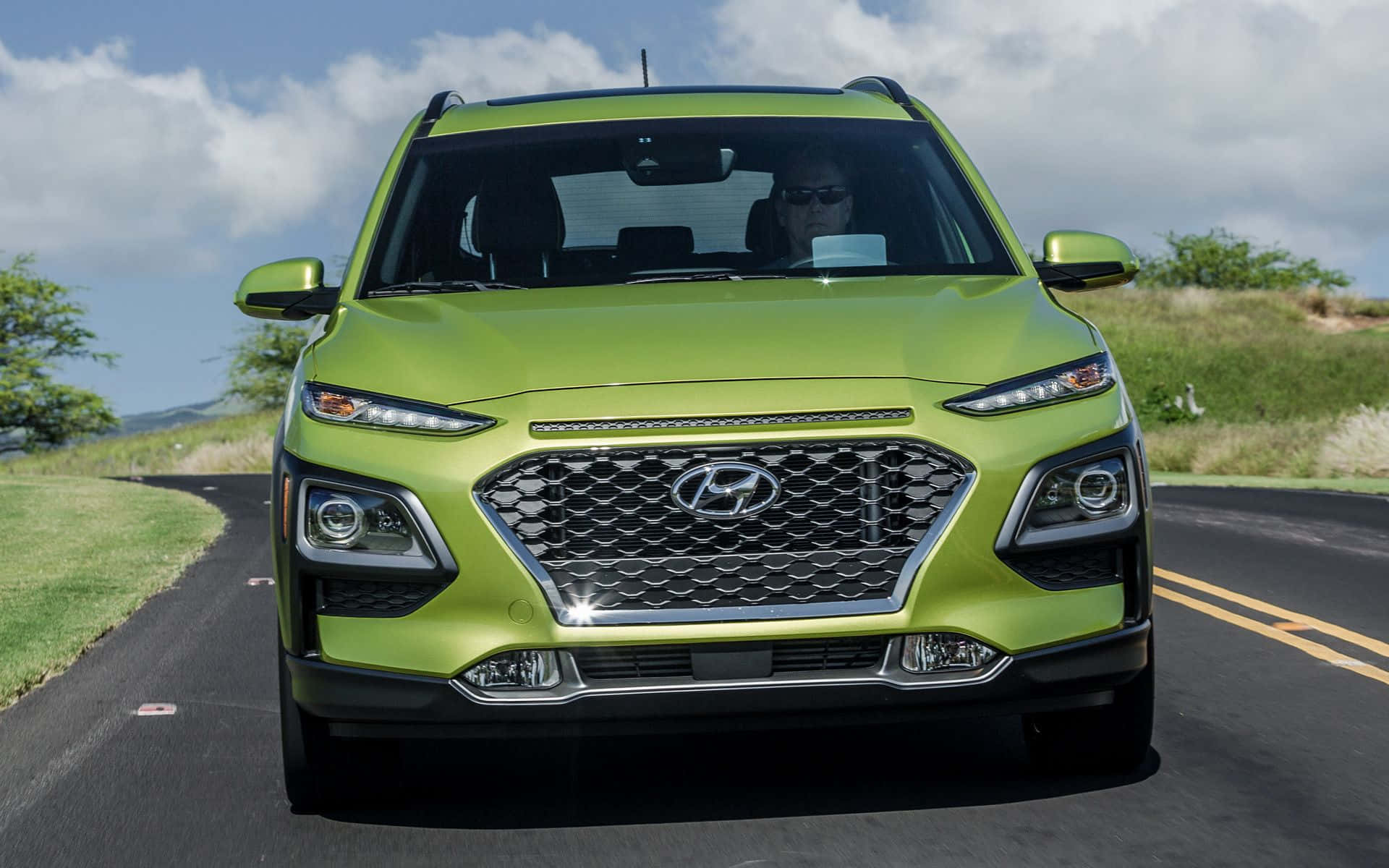 Hyundai Kona 桌布 壁紙 — 一輛充滿活力的萊姆綠色現代SUV在部分多雲的天空下沿著蜿蜒的道路正面行駛。
