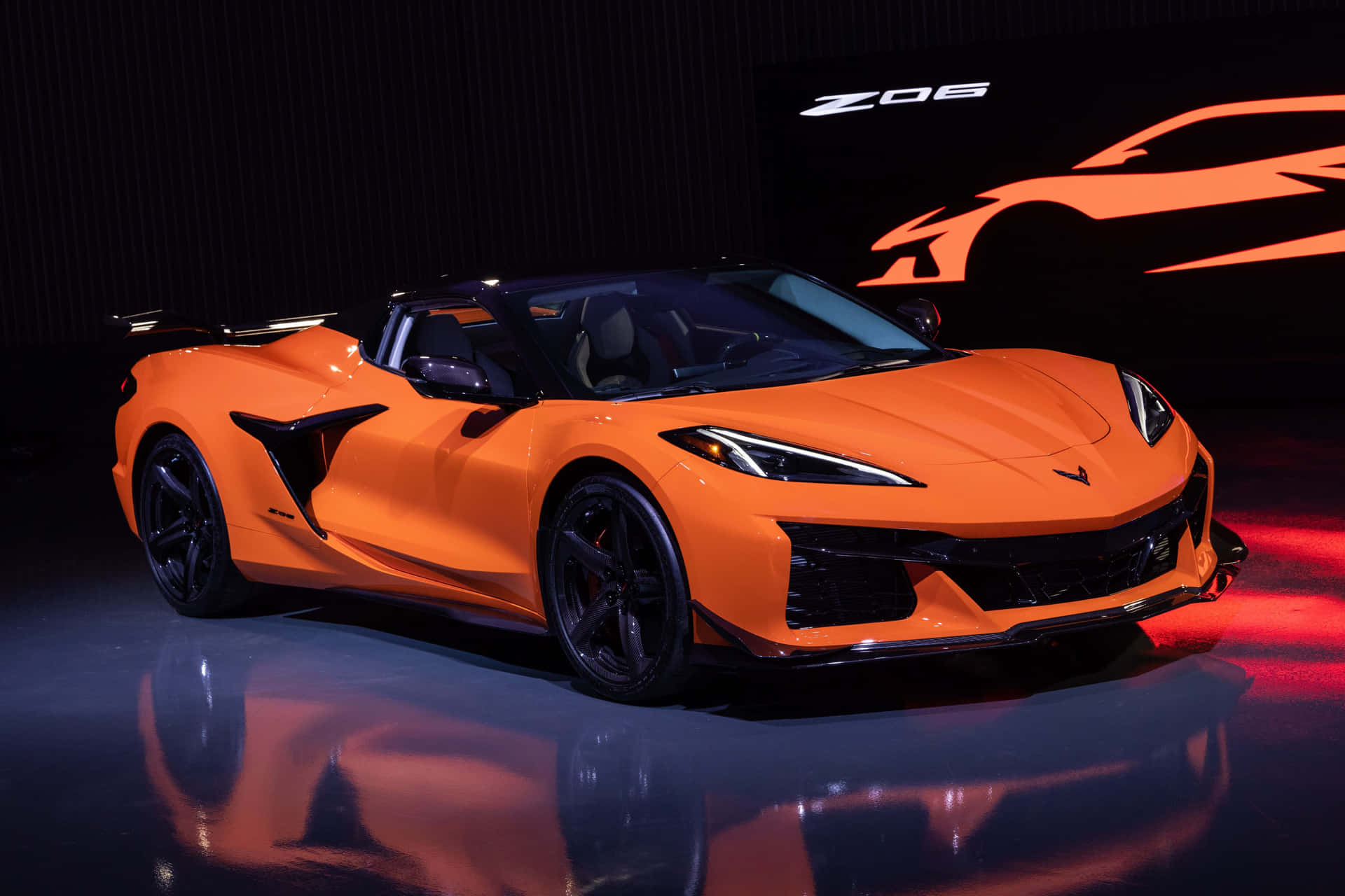  壁紙 — 一輛充滿活力的橙色Corvette Z06跑車在攝影棚燈光下，於反射舞台上閃耀，背景是發光的橙色剪影。