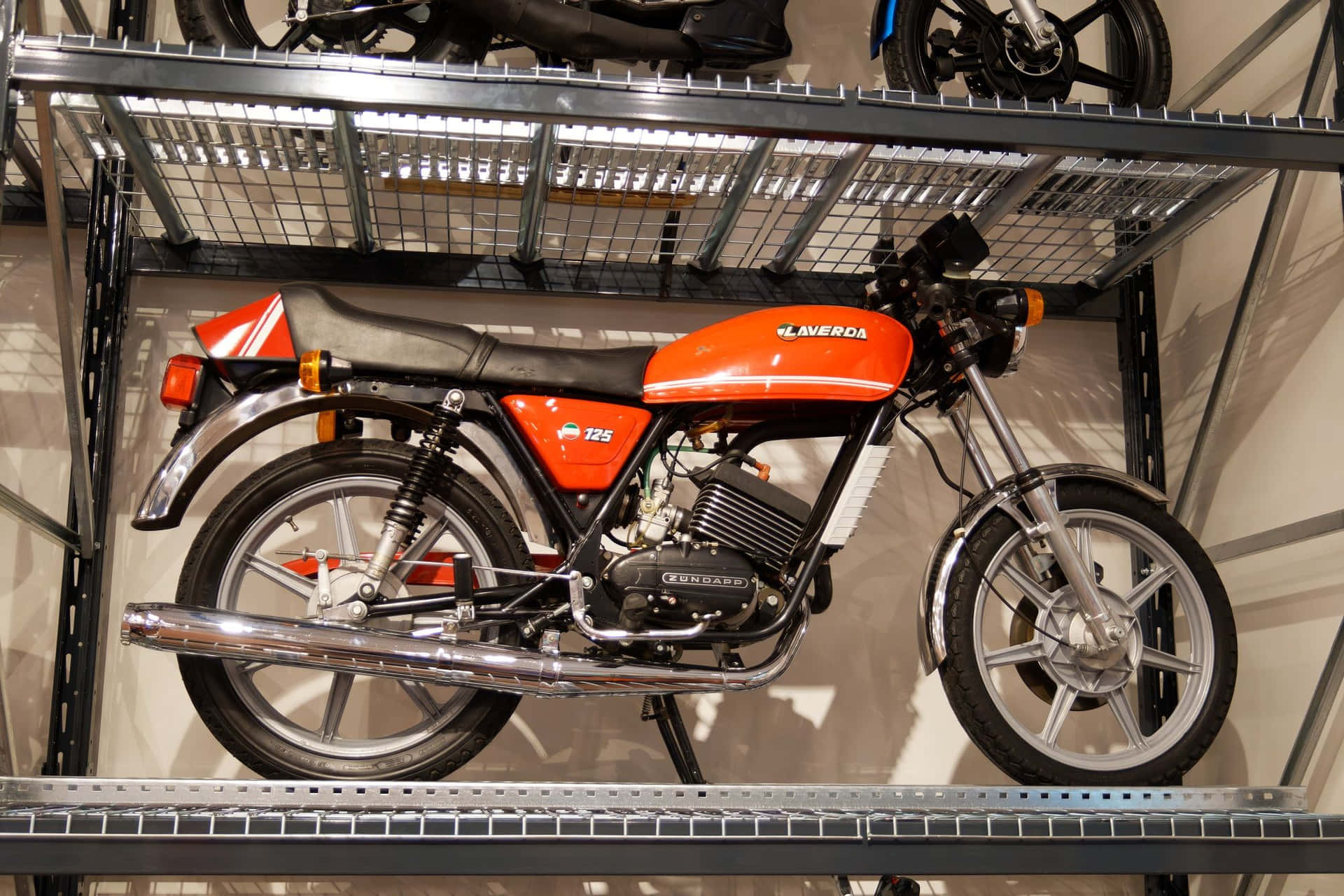  壁紙 — 一輛鮮豔的橙色Laverda 125摩托車展示在光線充足的室內車庫的金屬架上。