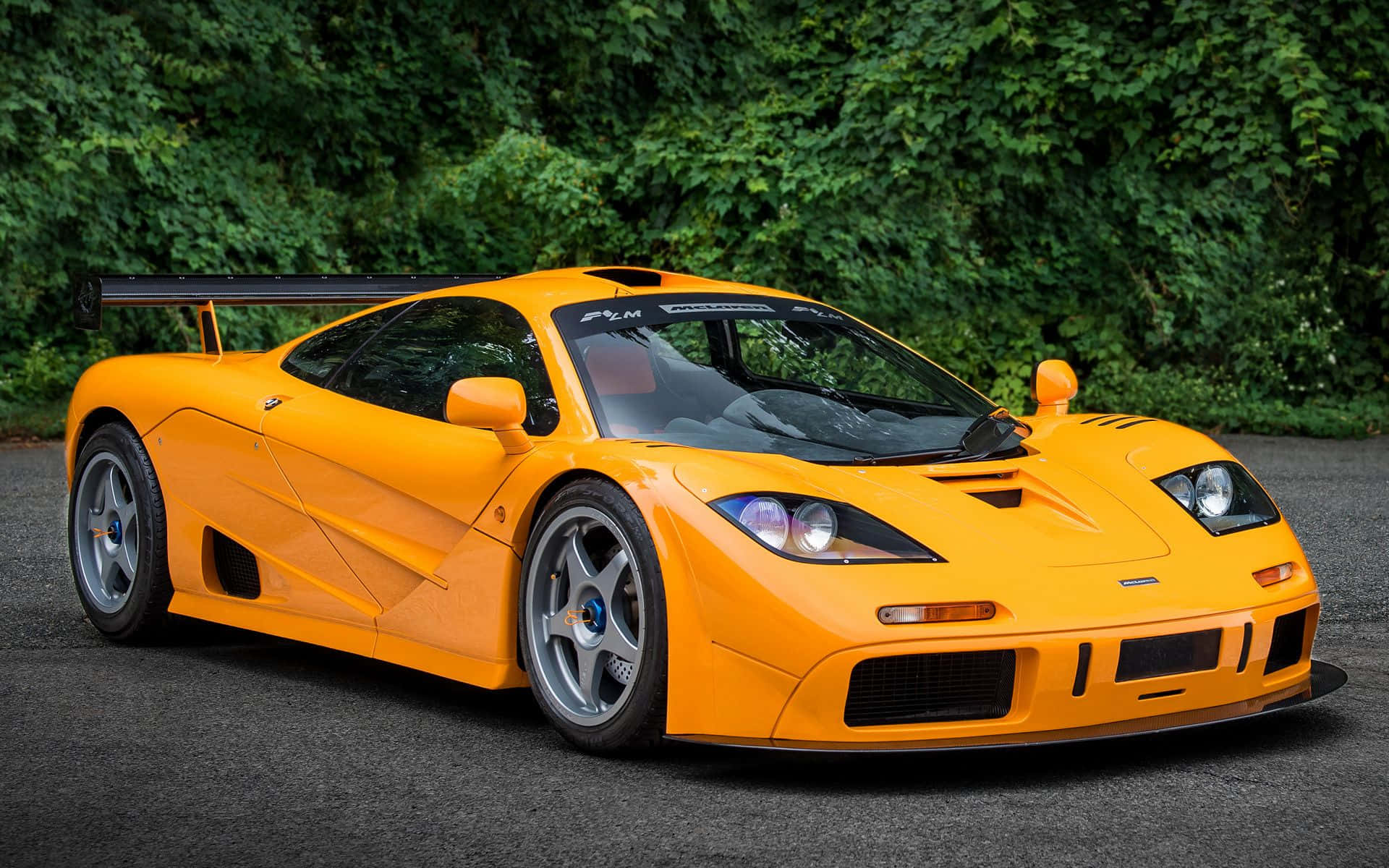 Mclaren F1 桌布 壁紙 — 一輛充滿活力的橙色McLaren F1 GTR跑車停在瀝青路上，背景是茂密的綠色樹葉。