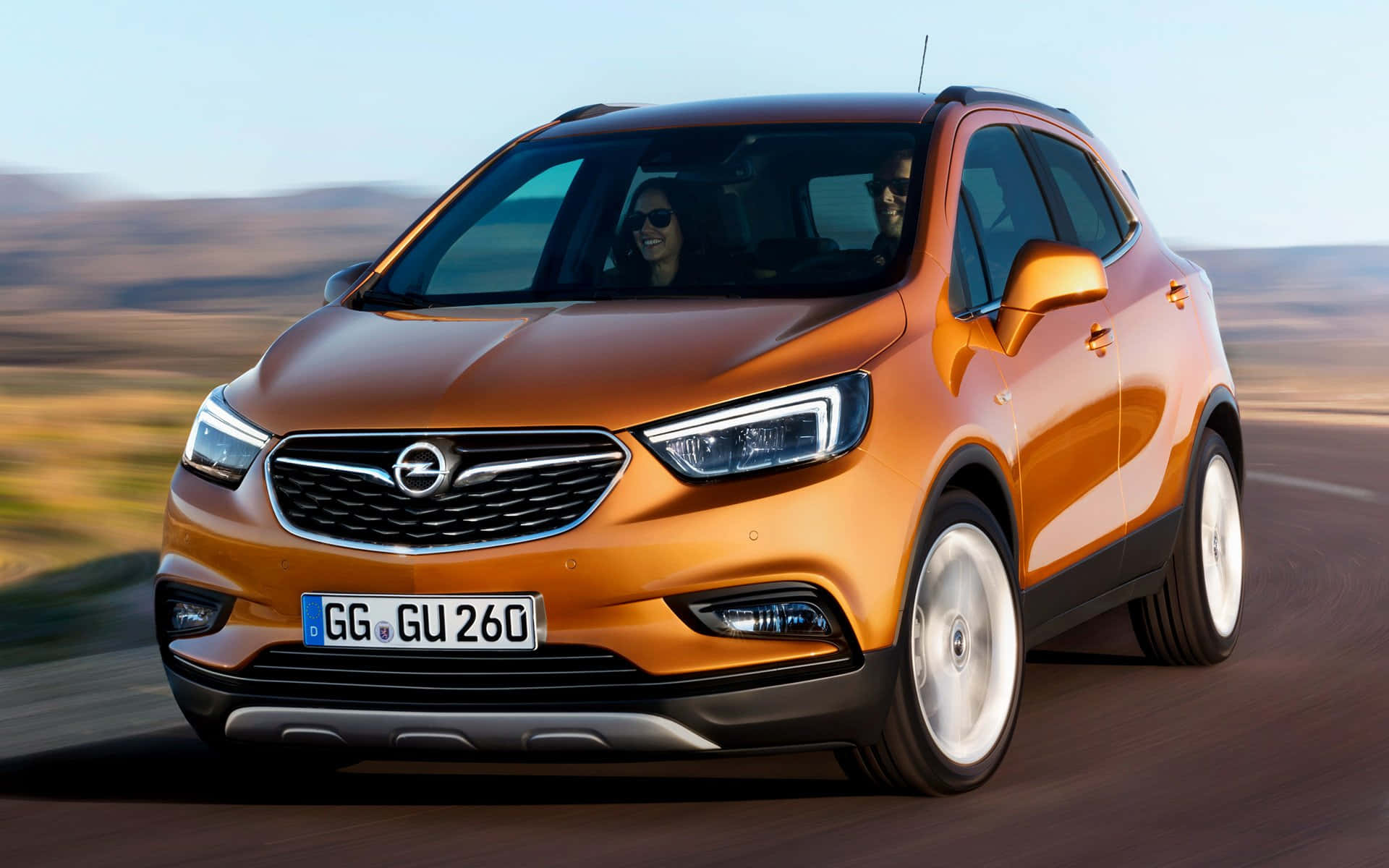 Opel Mokka 桌布 壁紙 — 一輛充滿活力的橙色Opel SUV在沙漠公路上疾馳，背景模糊，乘客面帶微笑。