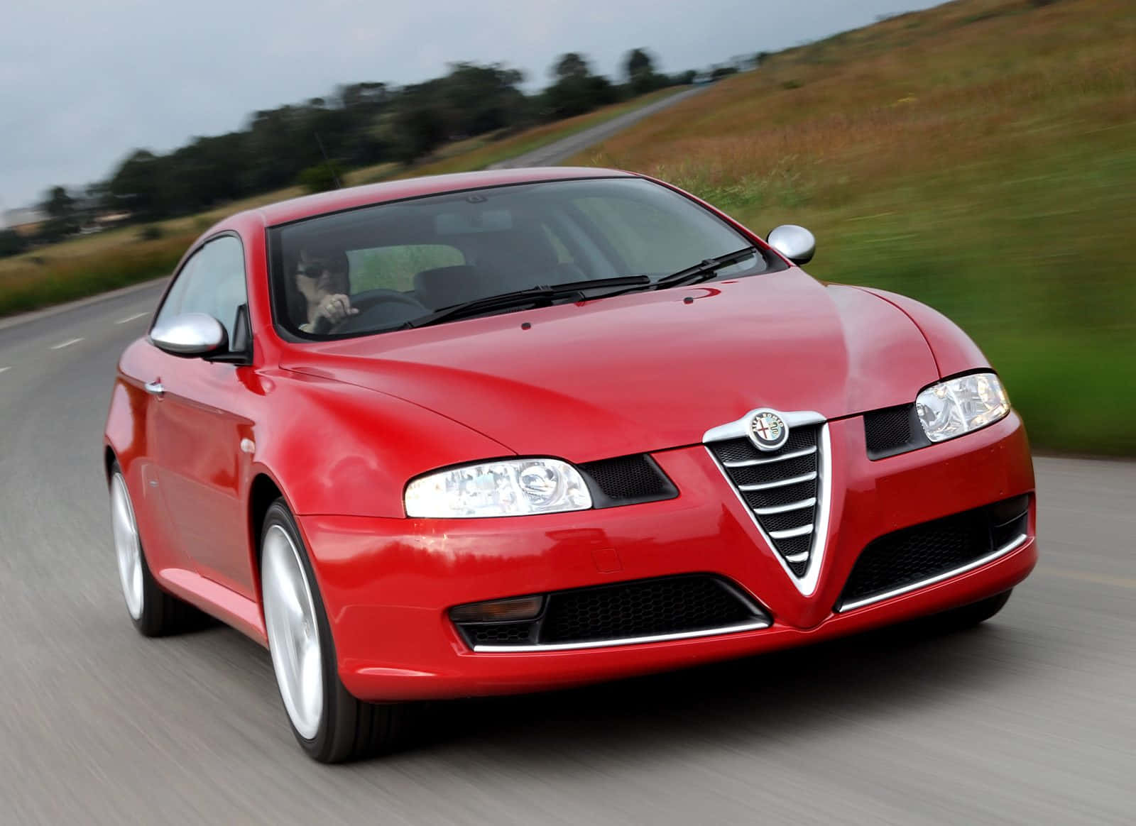 Alfa Romeo Gt 桌布 壁紙 — 一輛鮮紅色的阿爾法羅密歐跑車在蜿蜒的鄉村道路上疾馳，背景是模糊的綠色山丘。