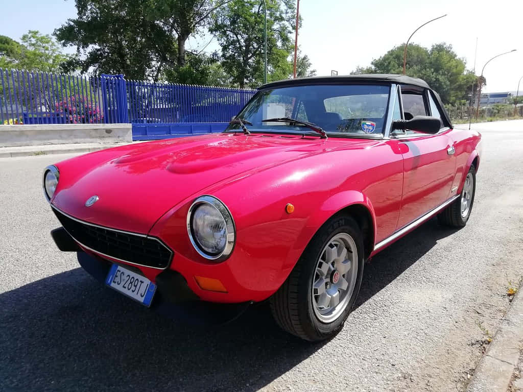 Fiat 124 Spider 桌布 壁紙 — 一輛鮮紅色的經典菲亞特蜘蛛敞篷車停泊在陽光照耀的瀝青路上，背景有樹木和藍色圍欄。