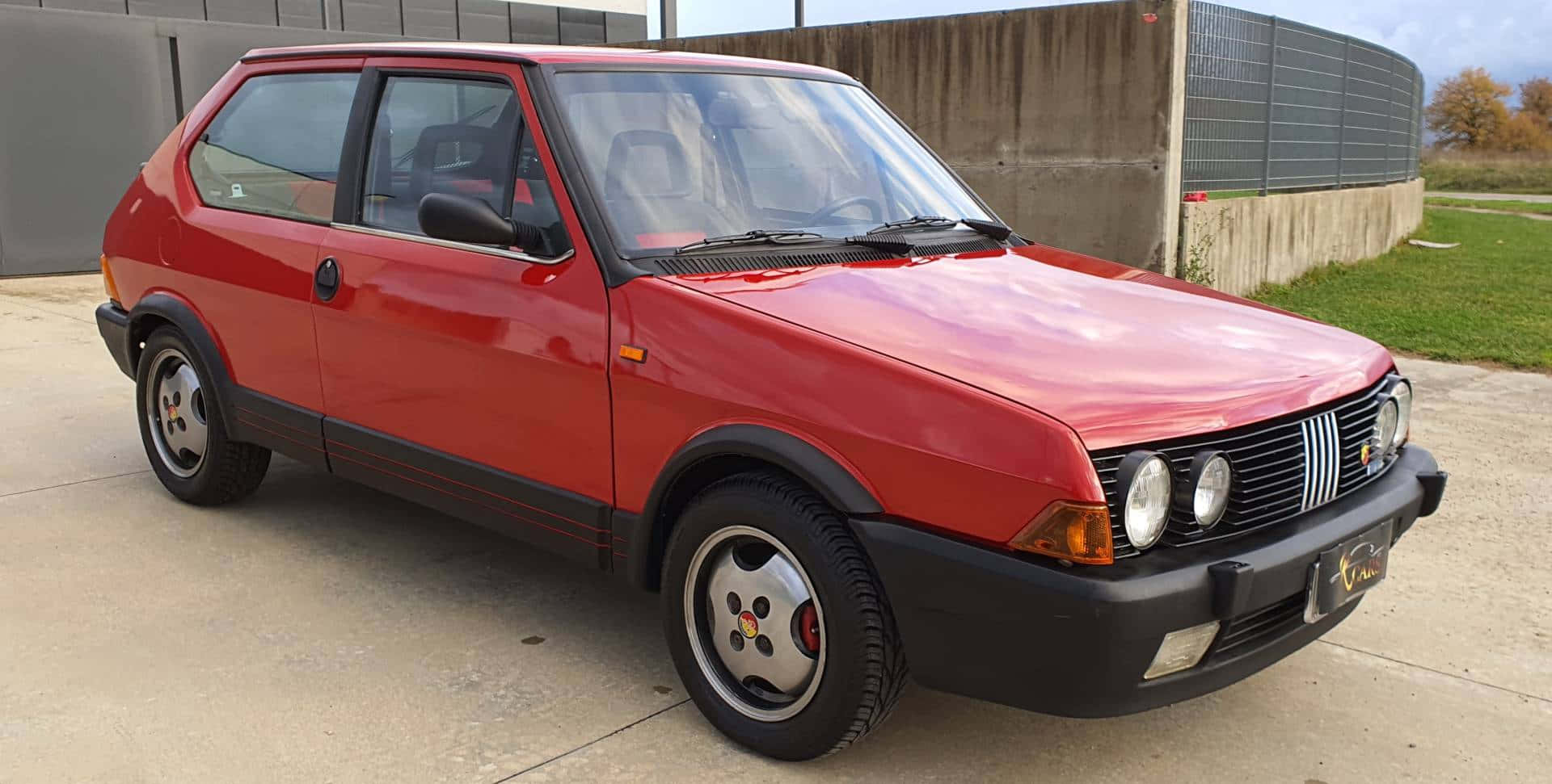 Fiat Ritmo 桌布 壁紙 — 一輛鮮紅色的經典飛雅特Uno停放在混凝土空地旁，旁邊有草地和混凝土牆。