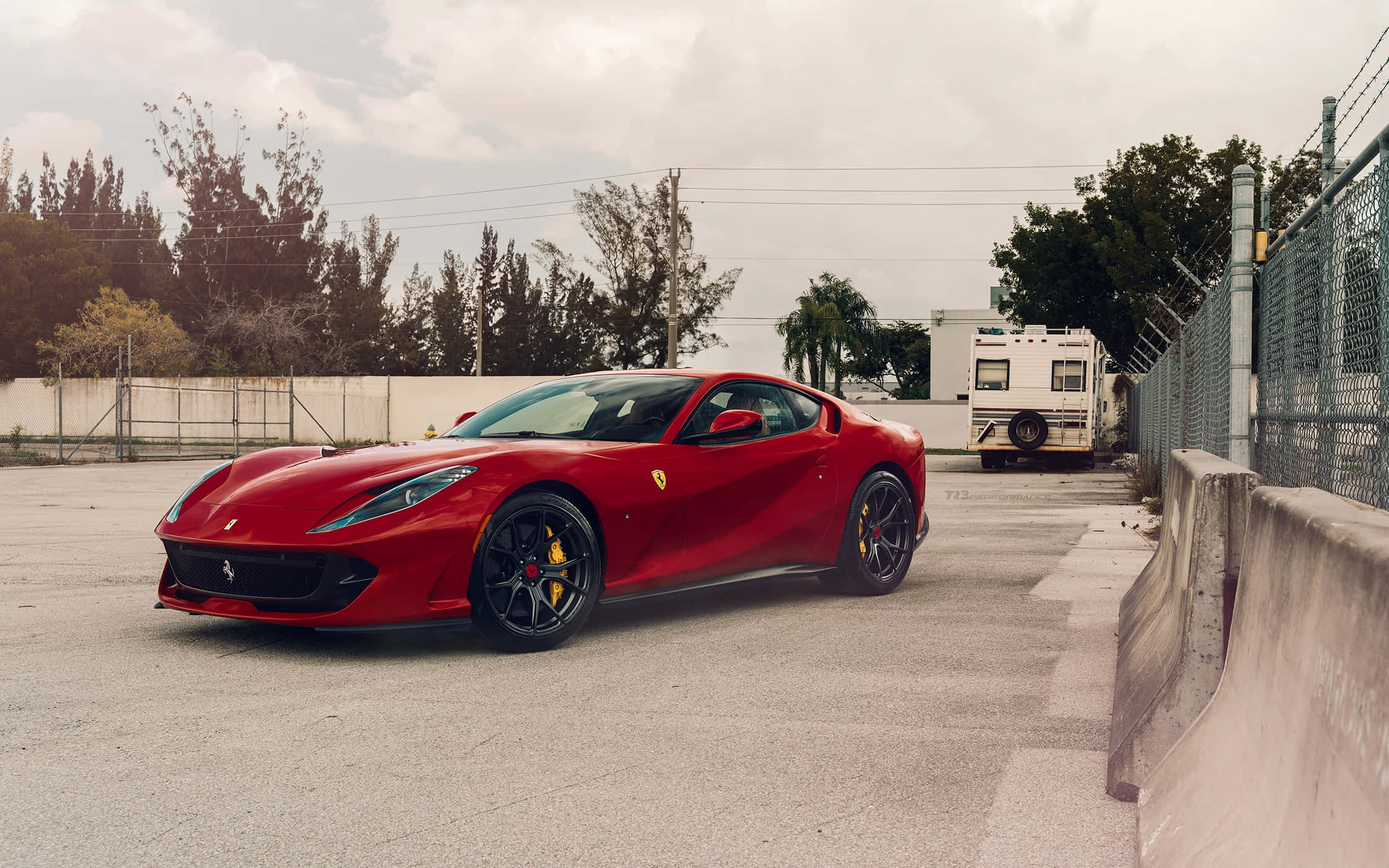 Ferrari 812 Superfast 桌布 壁紙 — 一輛鮮紅色的法拉利跑車停在混凝土場地上，旁邊是鏈條圍欄，背景有樹木和拖車。