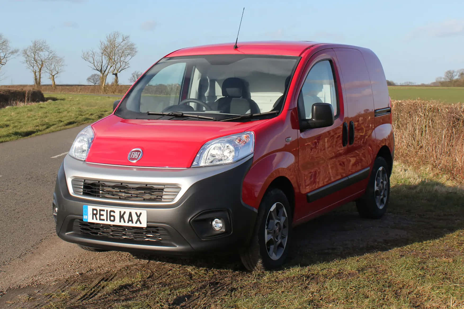 Fiat Fiorino 桌布 壁紙 — 一輛鮮紅色的Fiat廂型車停在鄉間路邊，周圍有光禿禿的樹木和開闊的田野，上方是晴朗的藍天。