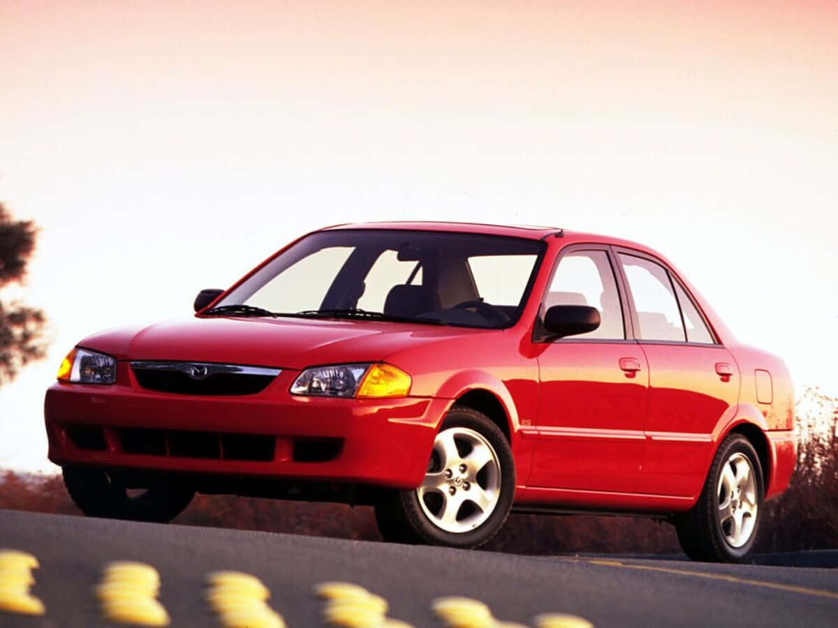 Mazda Protege 桌布 壁紙 — 一輛鮮紅色轎車在溫暖的夕陽光下閃閃發光，沿著帶有模糊黃色標記的路邊行駛。