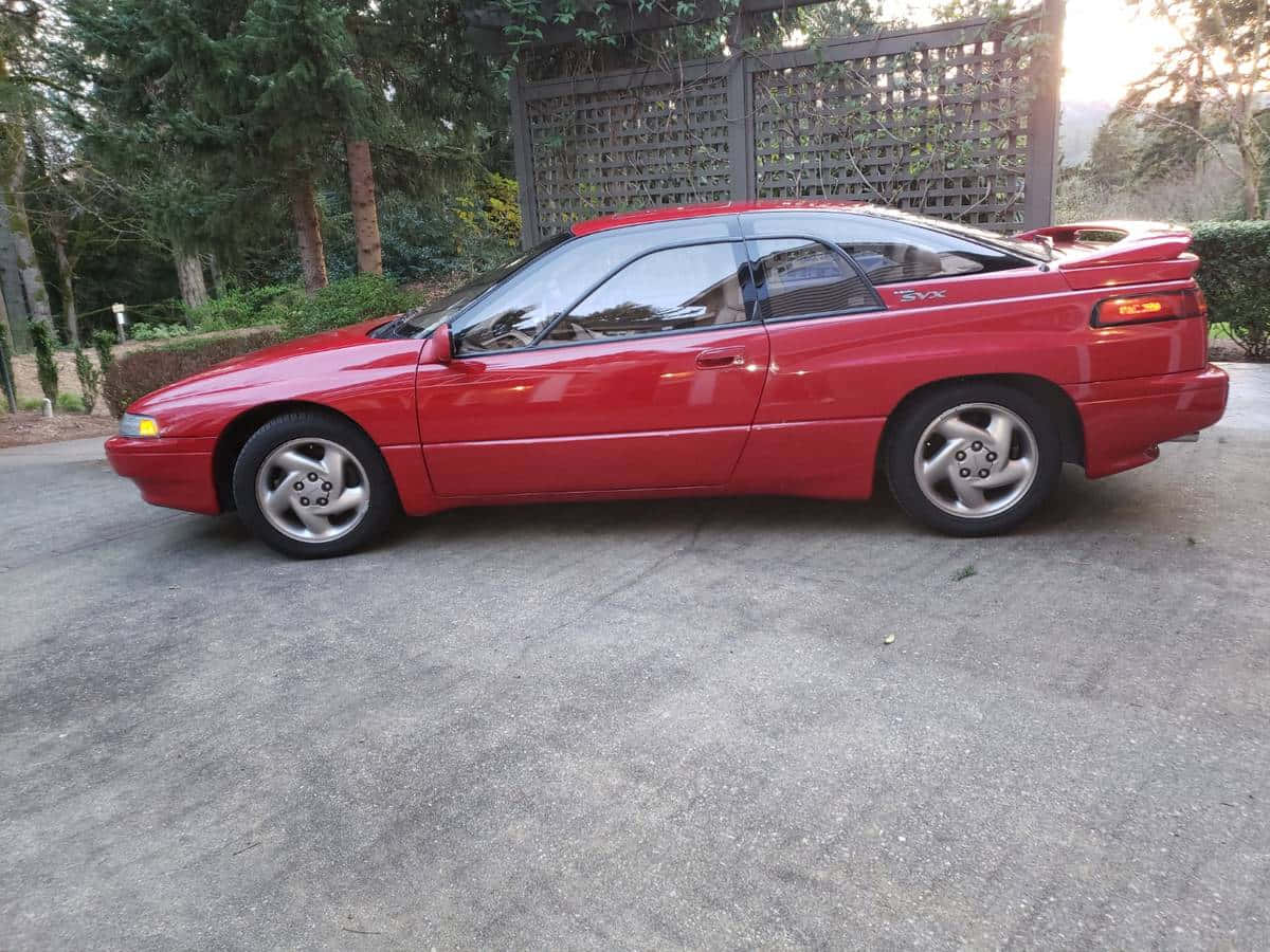 Subaru Svx 桌布 壁紙 — 一輛鮮紅色跑車停在混凝土車道上，背景有樹木和木製格子圍欄。