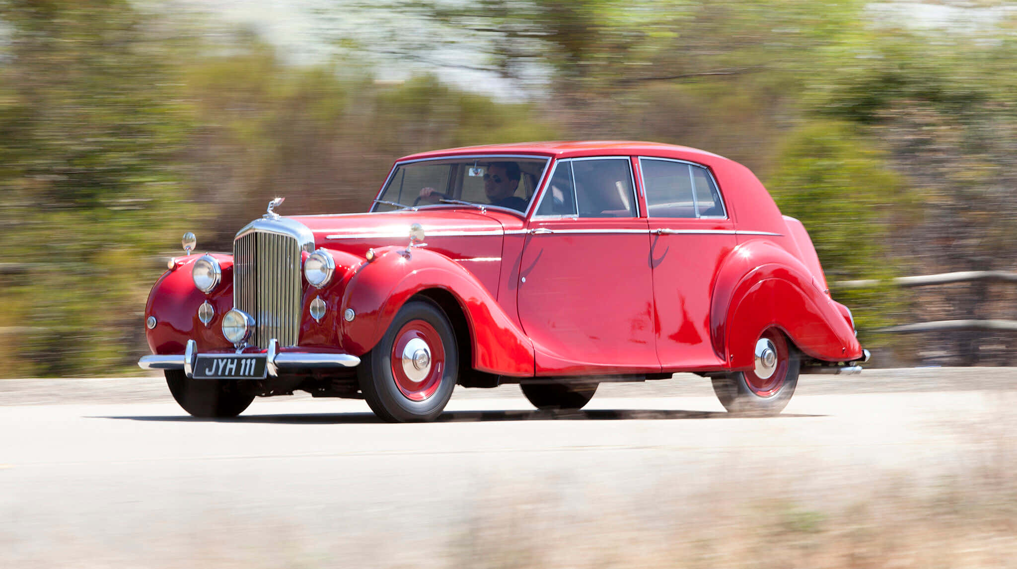 Bentley Mark Vi 桌布 壁紙 — 一輛鮮紅色的復古車在陽光明媚的道路上行駛，模糊的綠色植物飛馳而過。