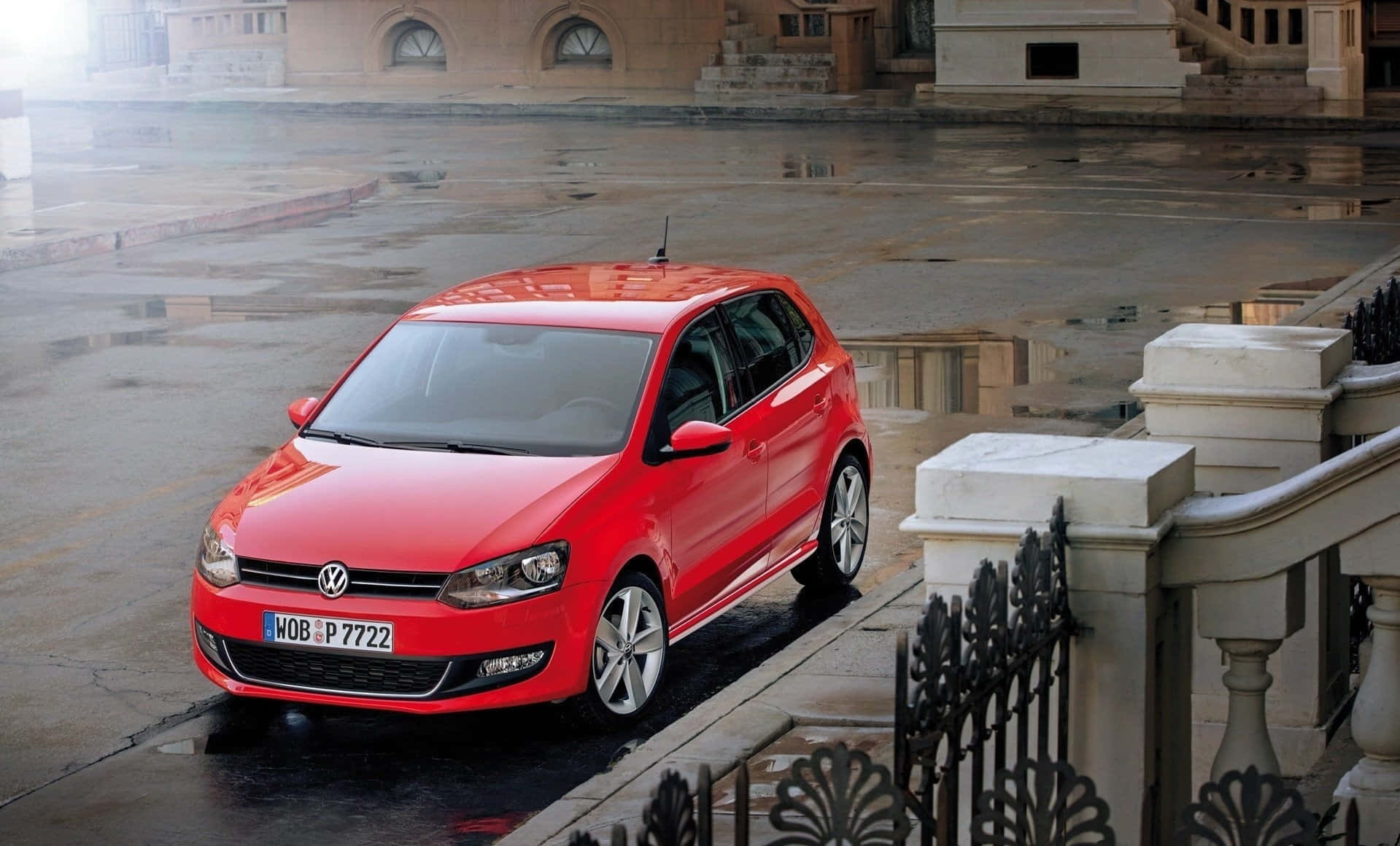 Volkswagen Polo 桌布 壁紙 — 一輛鮮紅色的福斯Polo停泊在濕漉漉的城市街道上，映照著多雲的天空和石頭建築。