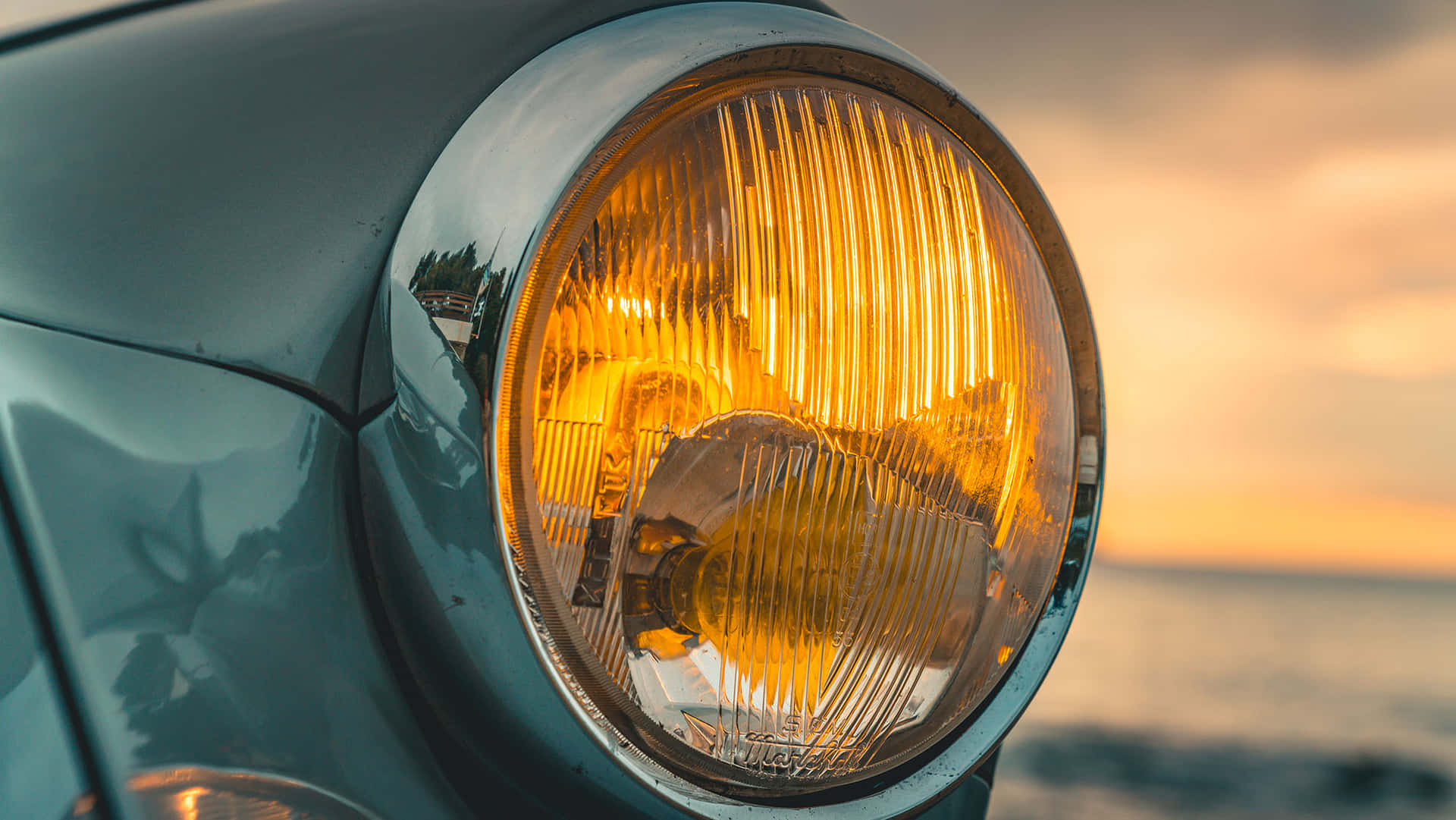  壁紙 — 一輛復古車的琥珀色車頭燈在水面上反射出夕陽，帶有溫暖的金色色調。