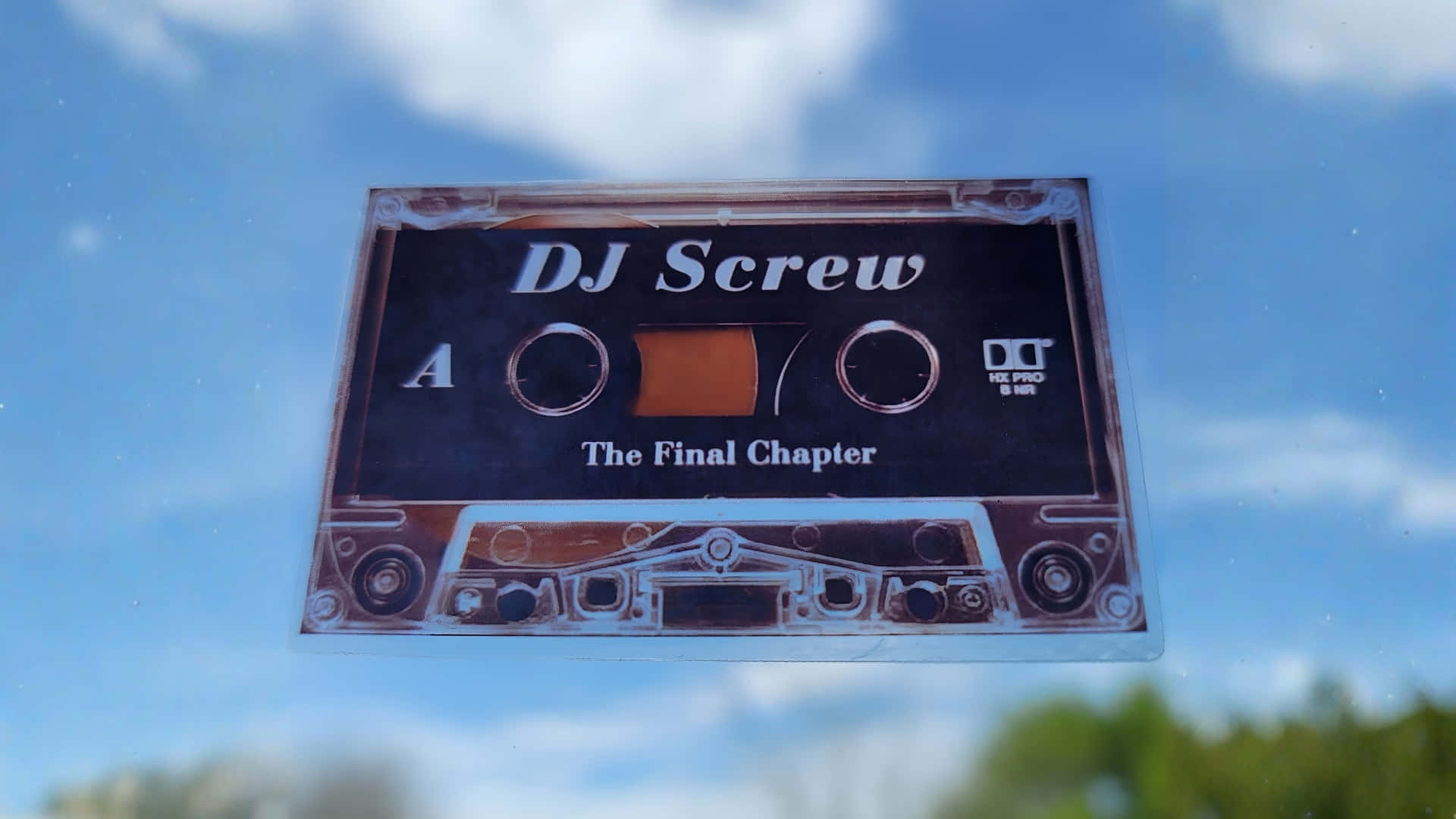  壁紙 — 一張復古的DJ Screw卡式錄音帶貼紙，上面有'The Final Chapter'文字，在明亮的藍天和蓬鬆的雲朵中反射。