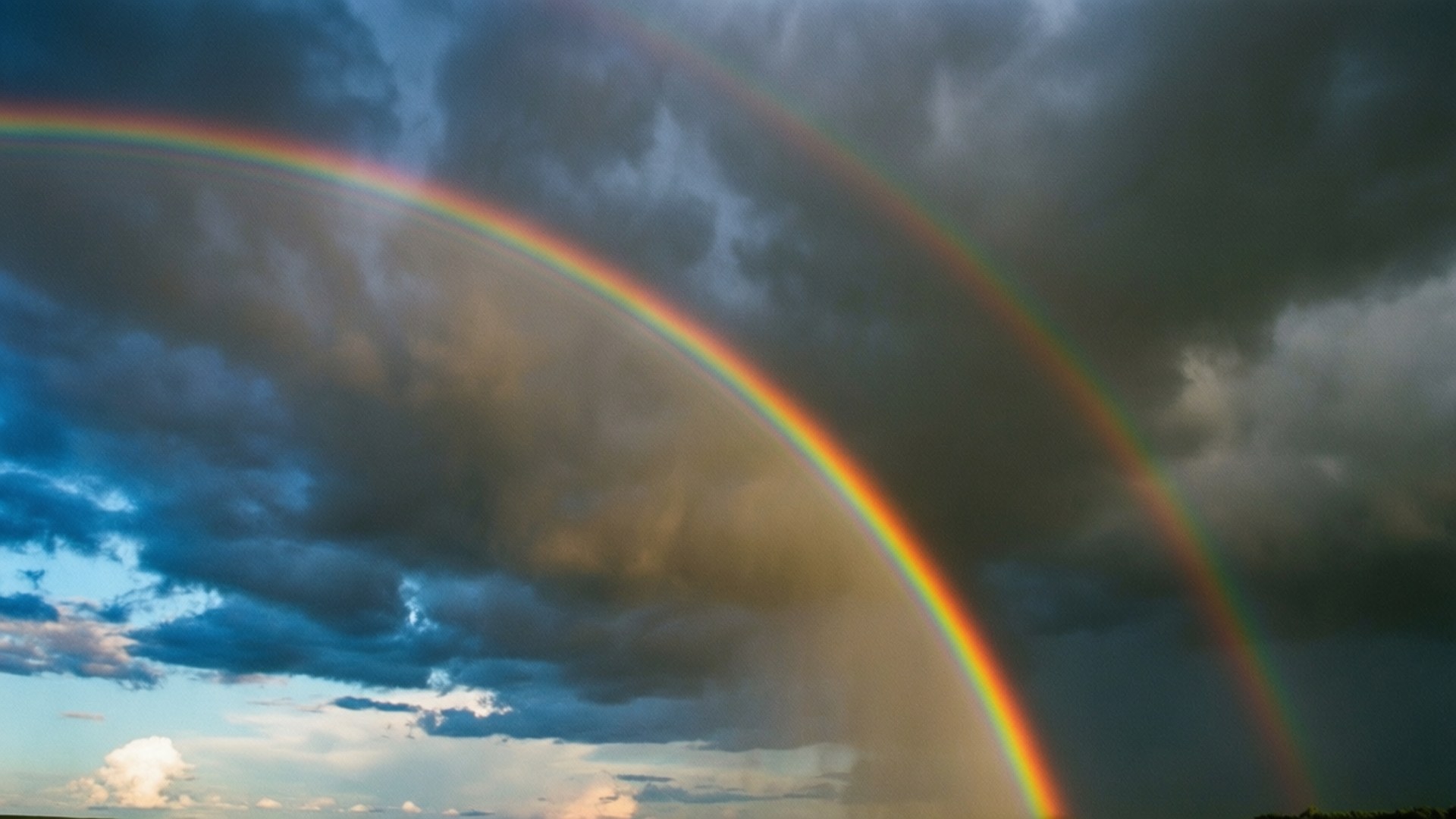 Double Rainbow Storm 壁紙 — 一條鮮豔的雙彩虹橫跨充滿黑暗風暴雲和藍色斑塊的戲劇性天空。
