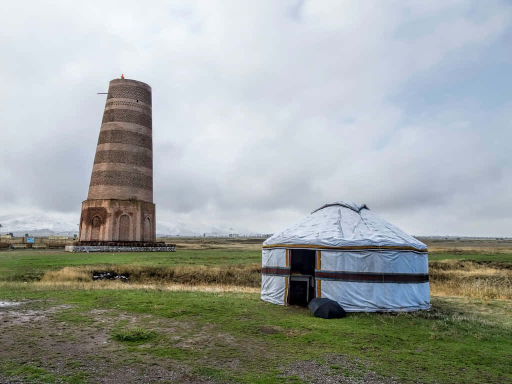 Kyrgyzstan 桌布 壁紙 — 一座風化的磚塔矗立在一片廣闊的草地上，旁邊有一個白色的蒙古包，天空多雲。