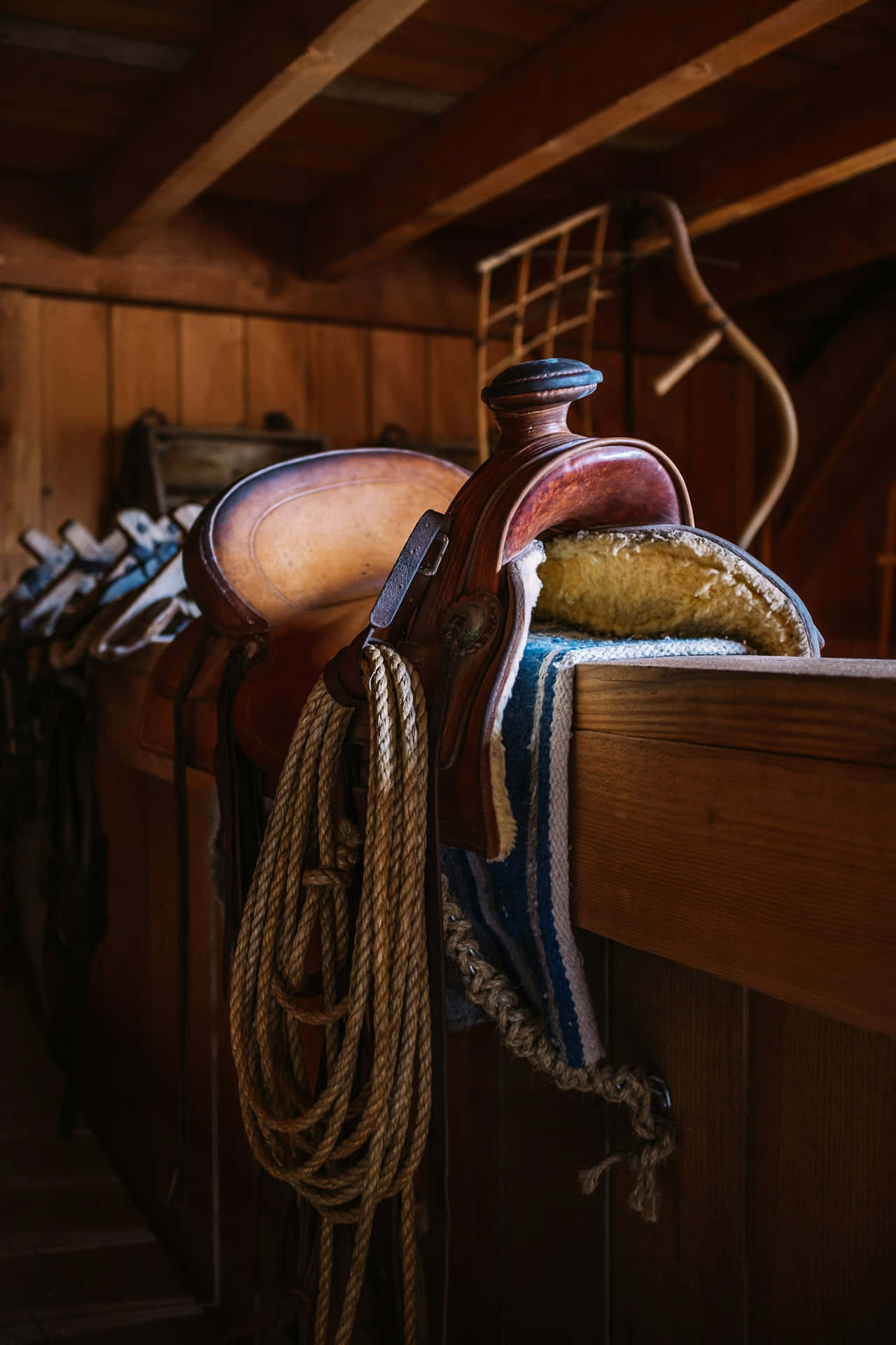  壁紙 — 一個風化的皮革馬鞍連繩索掛在一個有裸露橫樑的鄉村風格馬廄的木架上。
