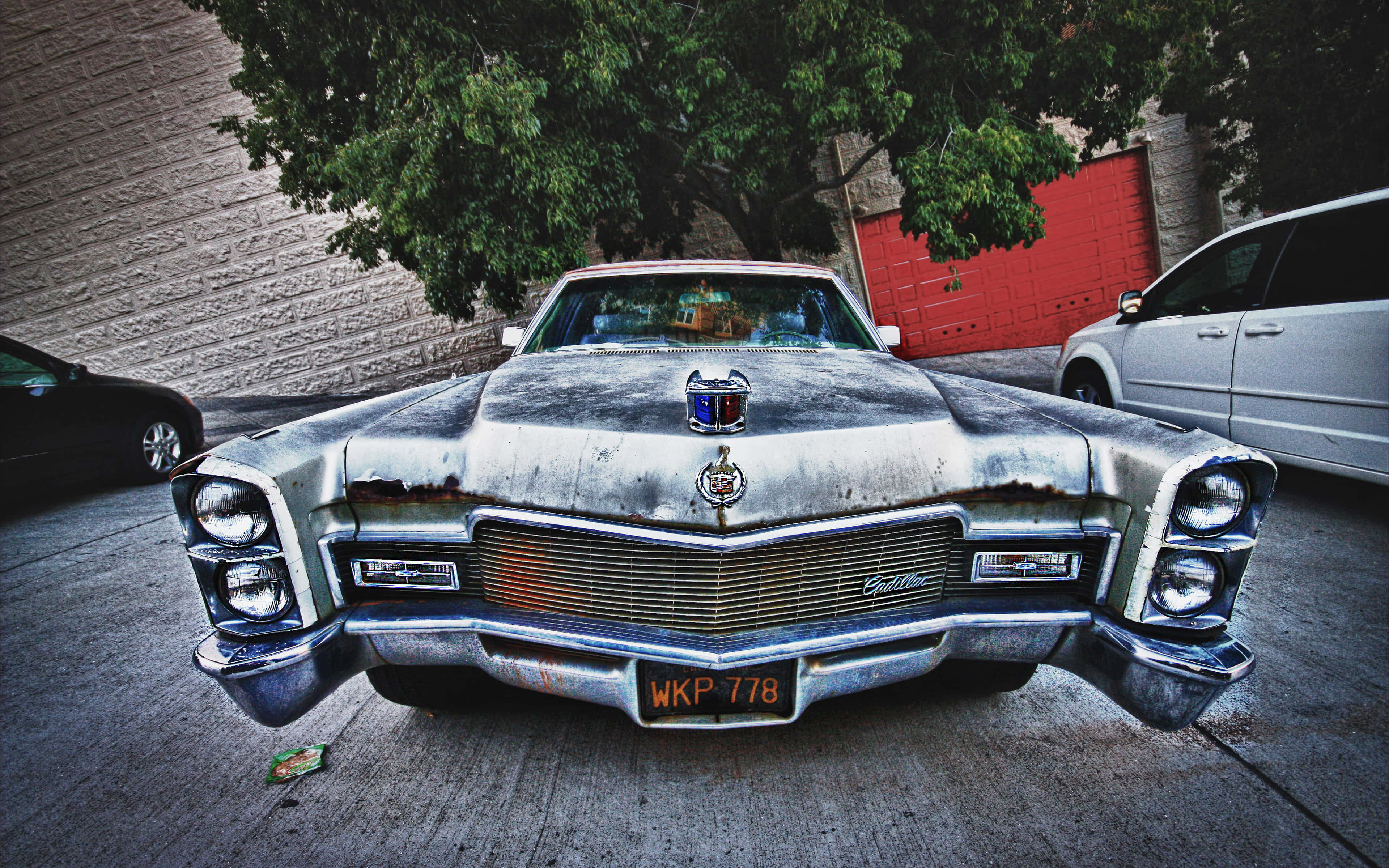 Cadillac Deville 桌布 壁紙 — 一輛風化的銀色經典凱迪拉克，帶有鍍鉻細節和鏽斑，停在樹下的混凝土街道上。