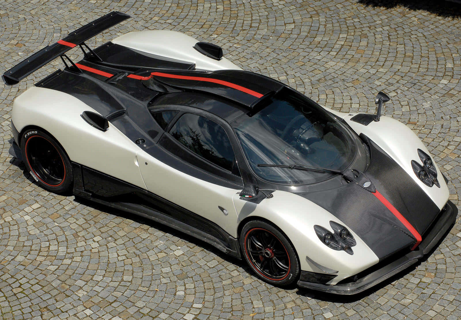Pagani Zonda Cinque 桌布 壁紙 — 一輛白色和黑色的Pagani超級跑車帶有紅色點綴，在明亮的日光下停放在彎曲的鵝卵石路面上。