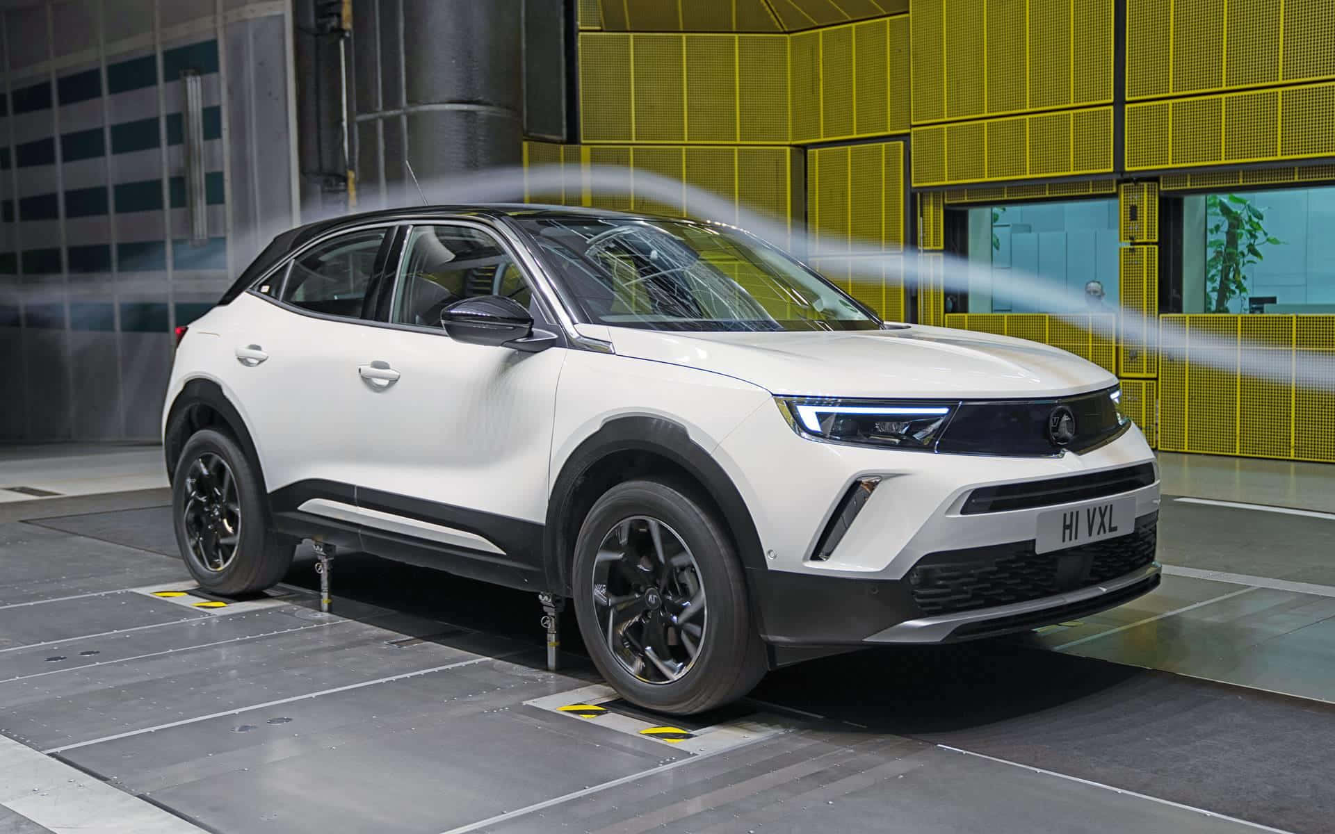 Opel Mokka 桌布 壁紙 — 一輛白色電動SUV在風洞中進行測試，可見氣流線條，背景為黃色和灰色的工業環境。