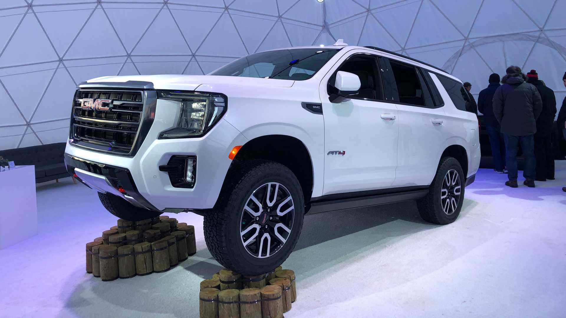 Gmc Yukon 桌布 壁紙 — 一輛白色GMC SUV被抬高在木頭上，位於未來風格的測地線穹頂展廳內，有觀察的人們。