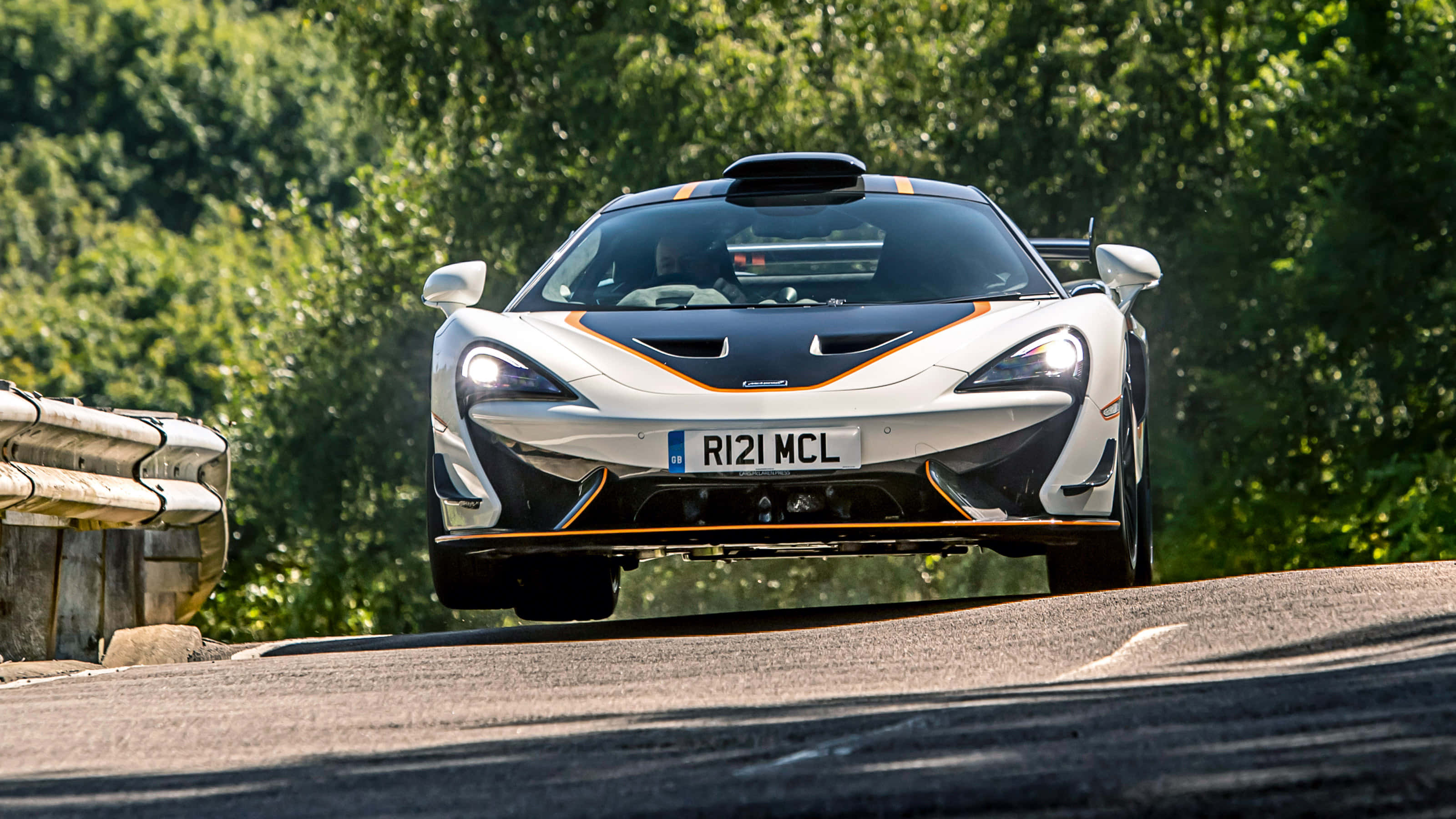 Mclaren 620r 桌布 壁紙 — 一輛帶有橙色飾邊的白色McLaren 570S在陽光明媚的森林道路上疾馳，車頭燈穿透綠意。