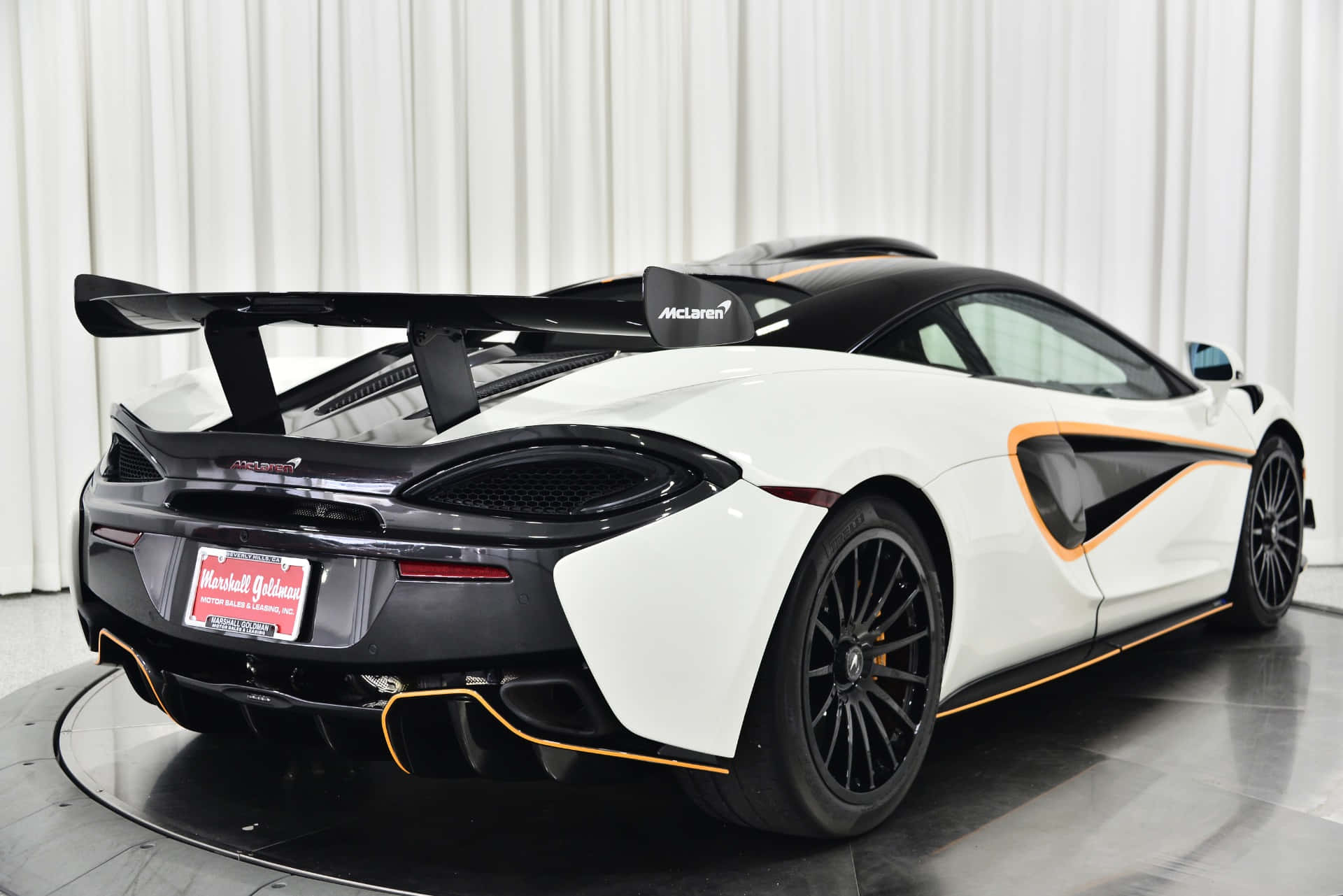 Mclaren 620r 桌布 壁紙 — 一輛白色McLaren跑車帶有黑色和橙色點綴，從後方四分之三角度在旋轉平台上展示，背景為白色窗簾。