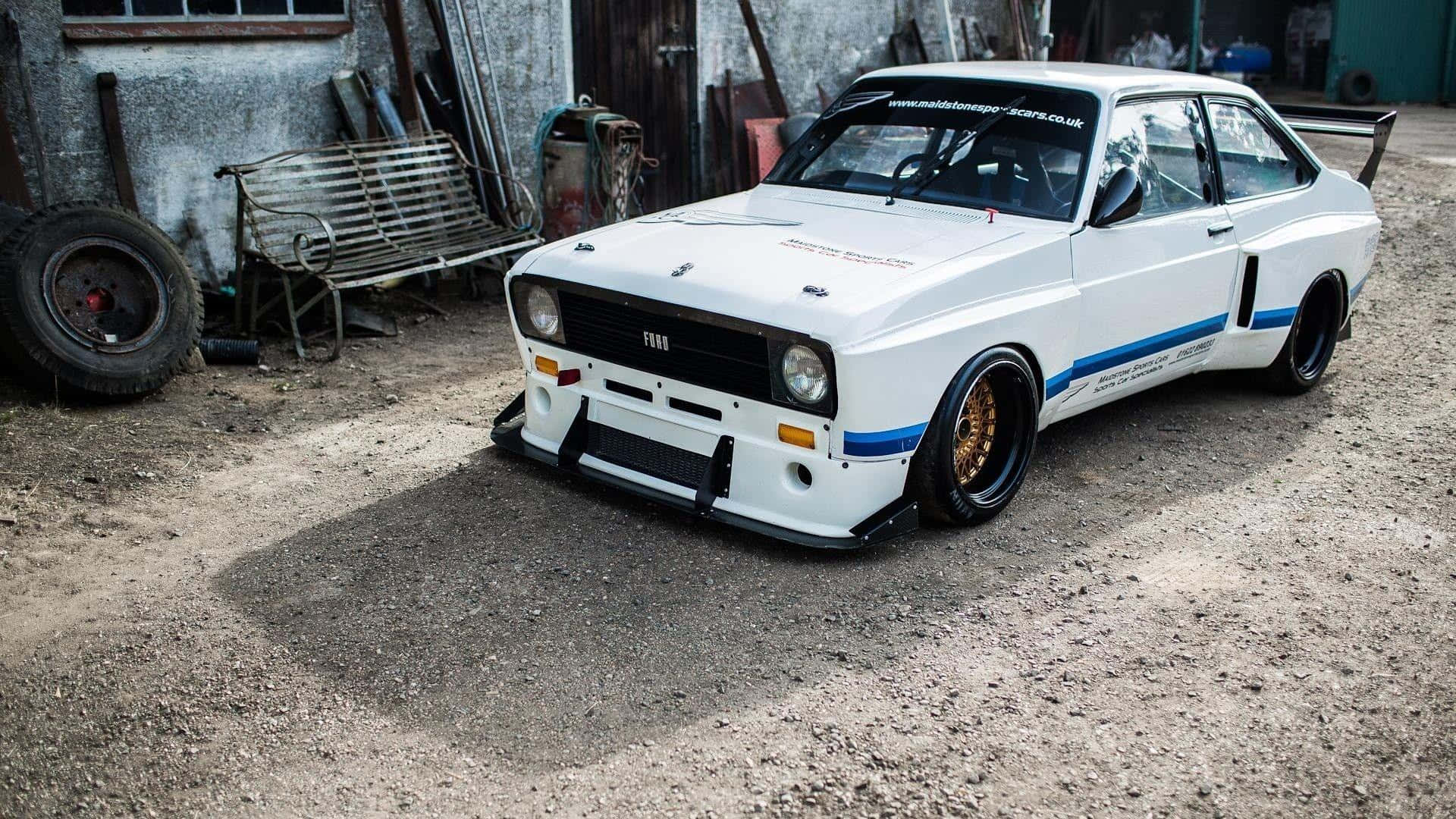 Ford Escort 桌布 壁紙 — 一輛白色改裝的Fiat拉力賽車，帶有藍色條紋和金色輪圈，停放在一個鄉村風格車庫庭院的碎石地上。