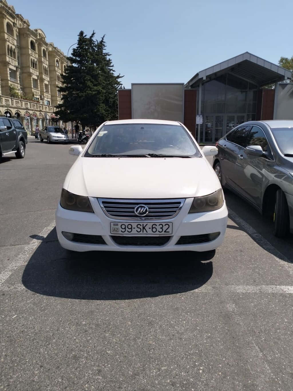  壁紙 — 一輛白色轎車停放在陽光明媚的城市地段，附近有歷史建築和其他汽車。
