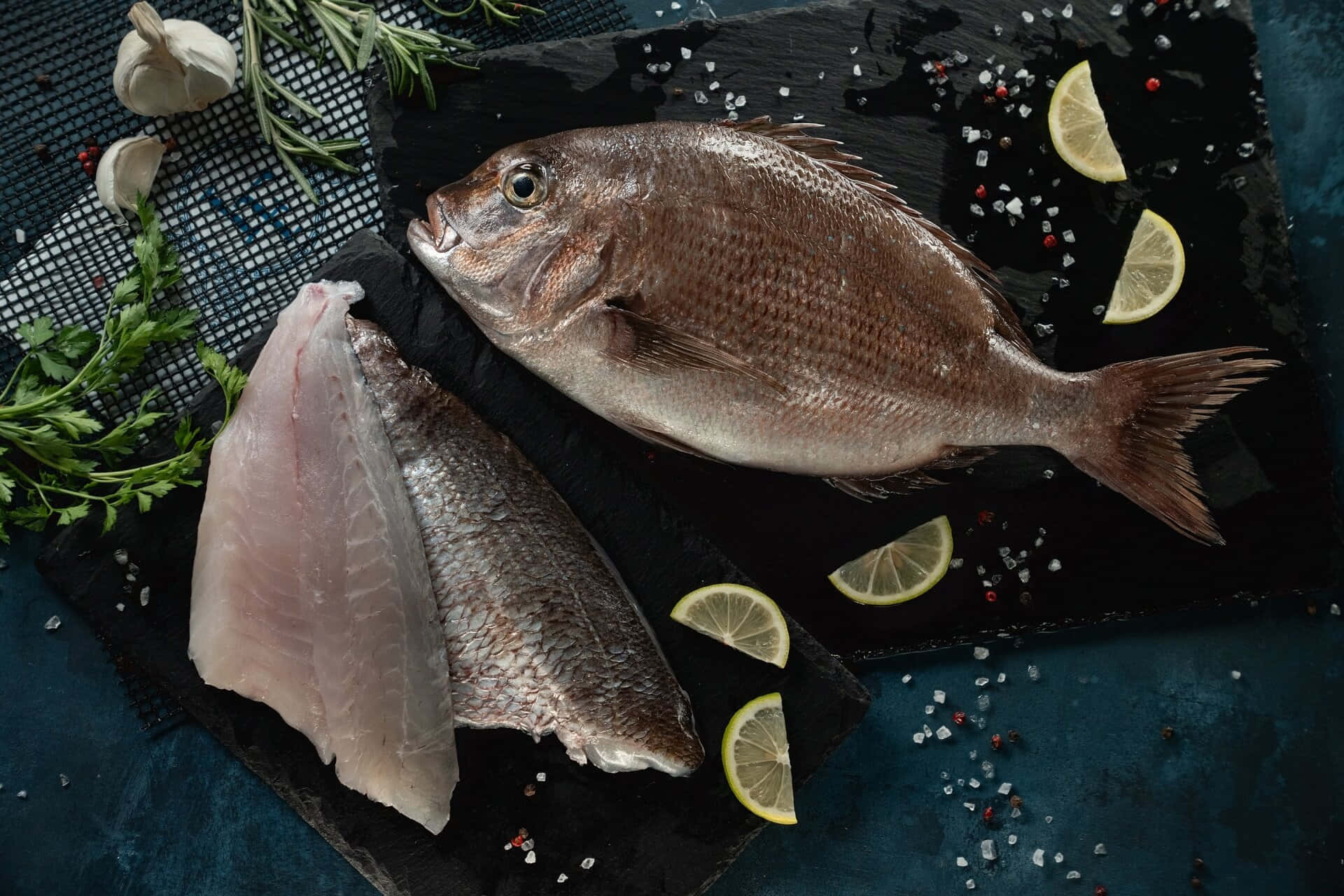  壁紙 — 一條完整的魚和魚片放在深色石板板上，配上檸檬片、大蒜、迷迭香和鹽，營造出質樸的烹飪場景。