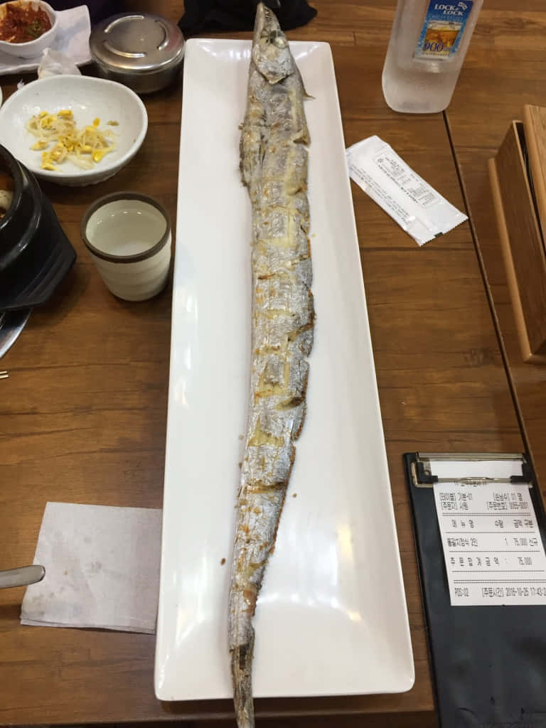  壁紙 — 一條烤全魚放在木桌上的白色盤子上，周圍是韓國小菜和飲料。