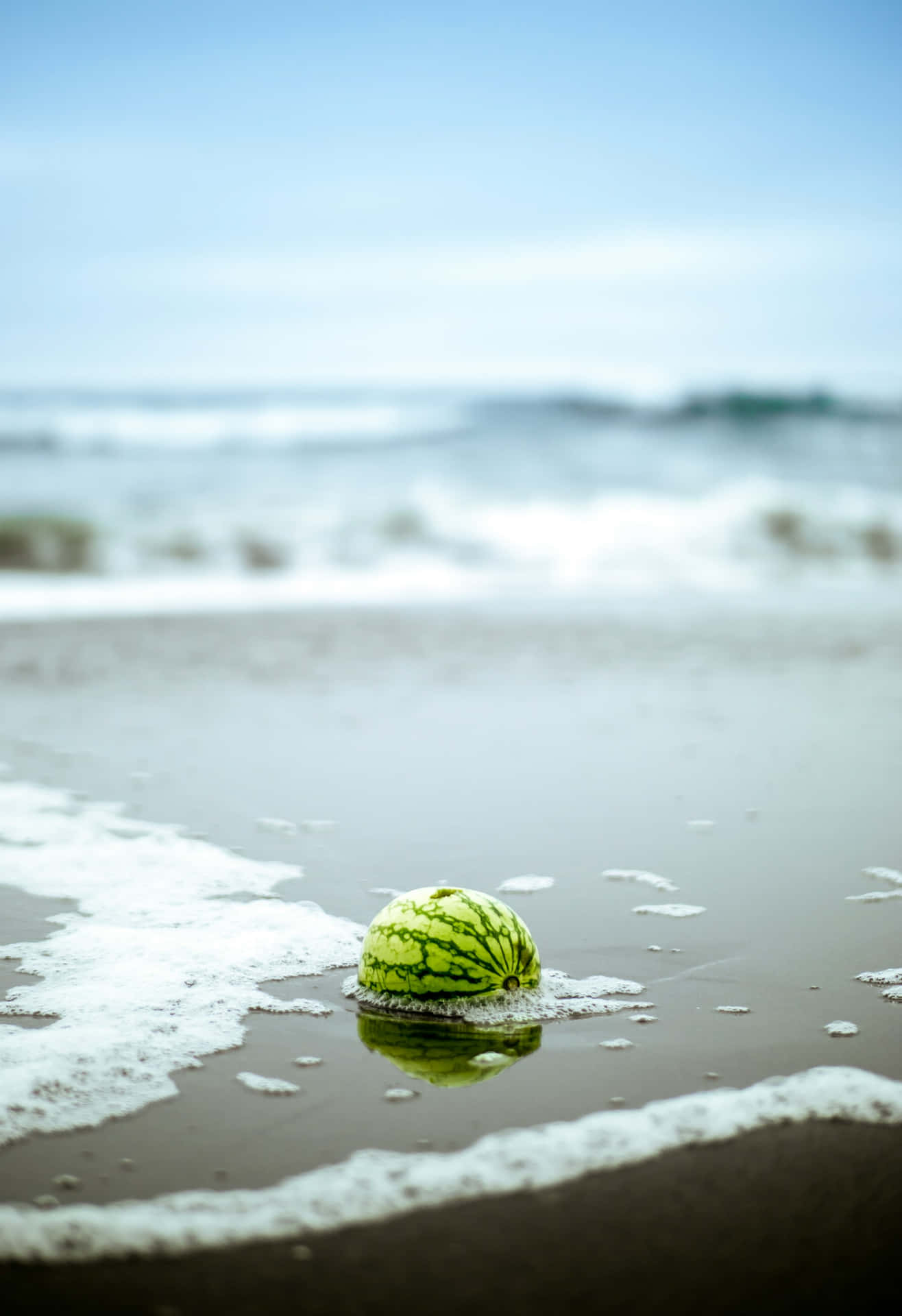  壁紙 — 一個完整的西瓜放在沙灘的淺海水面上，伴隨著溫柔的海浪和清澈的藍天。