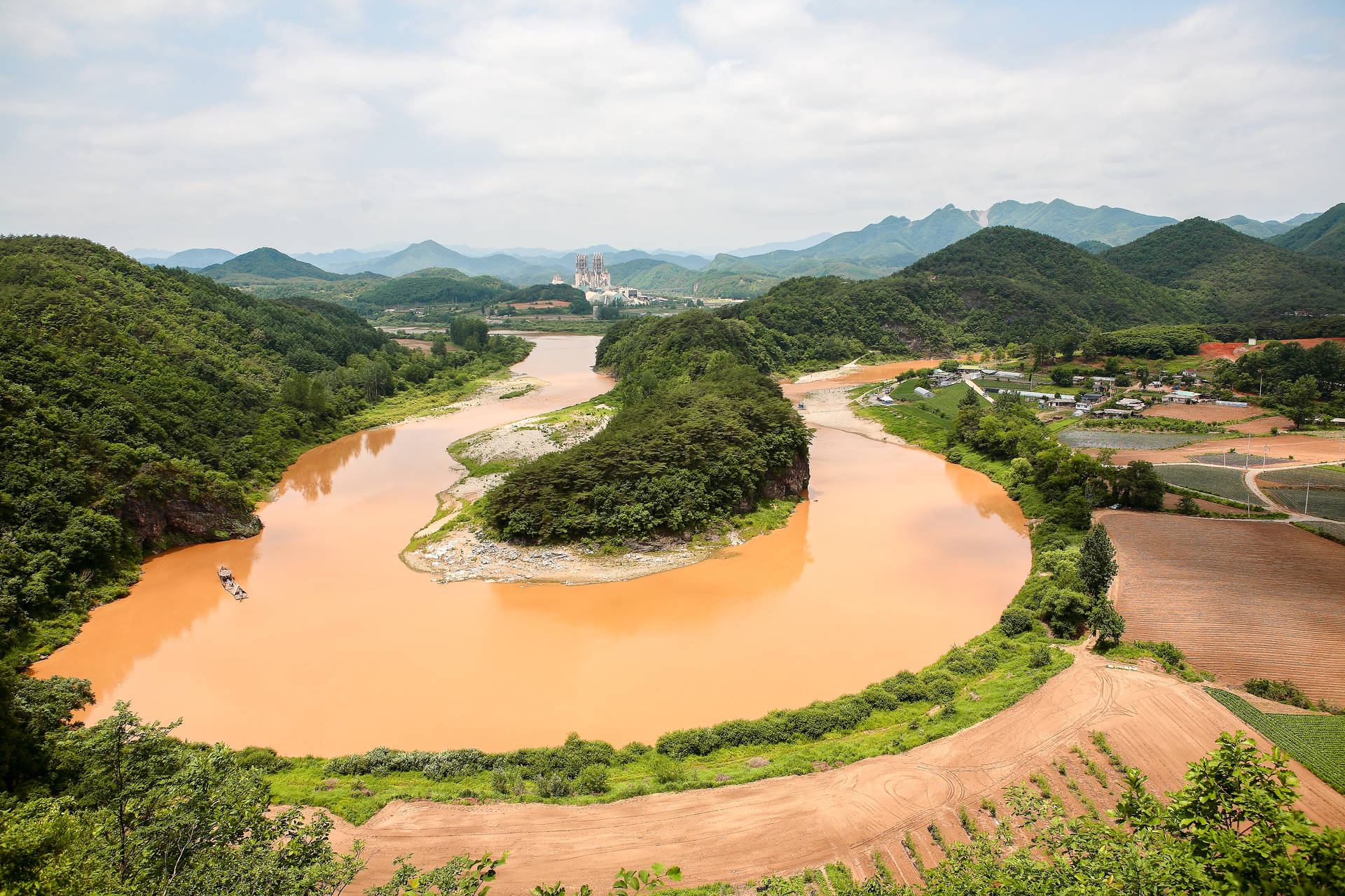 一條泥濘的河流蜿蜒穿過綠色山丘和農田，遠處有工廠。 — 韓劇 壁紙