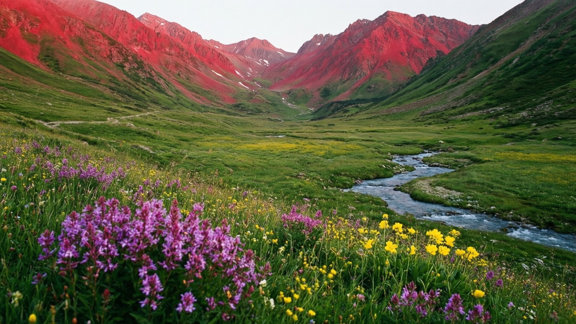 Flower Valley 壁紙 — 一條蜿蜒的小溪流過山谷，山谷中有綠草和紫黃色野花，下方是夕陽下的紅色山脈。