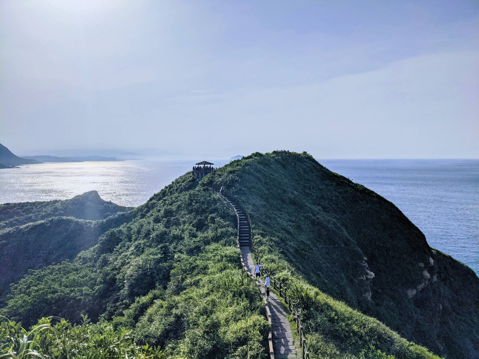 一條木棧道爬上翠綠的山丘，通往一個可欣賞海景的亭子。 — 台北 壁紙